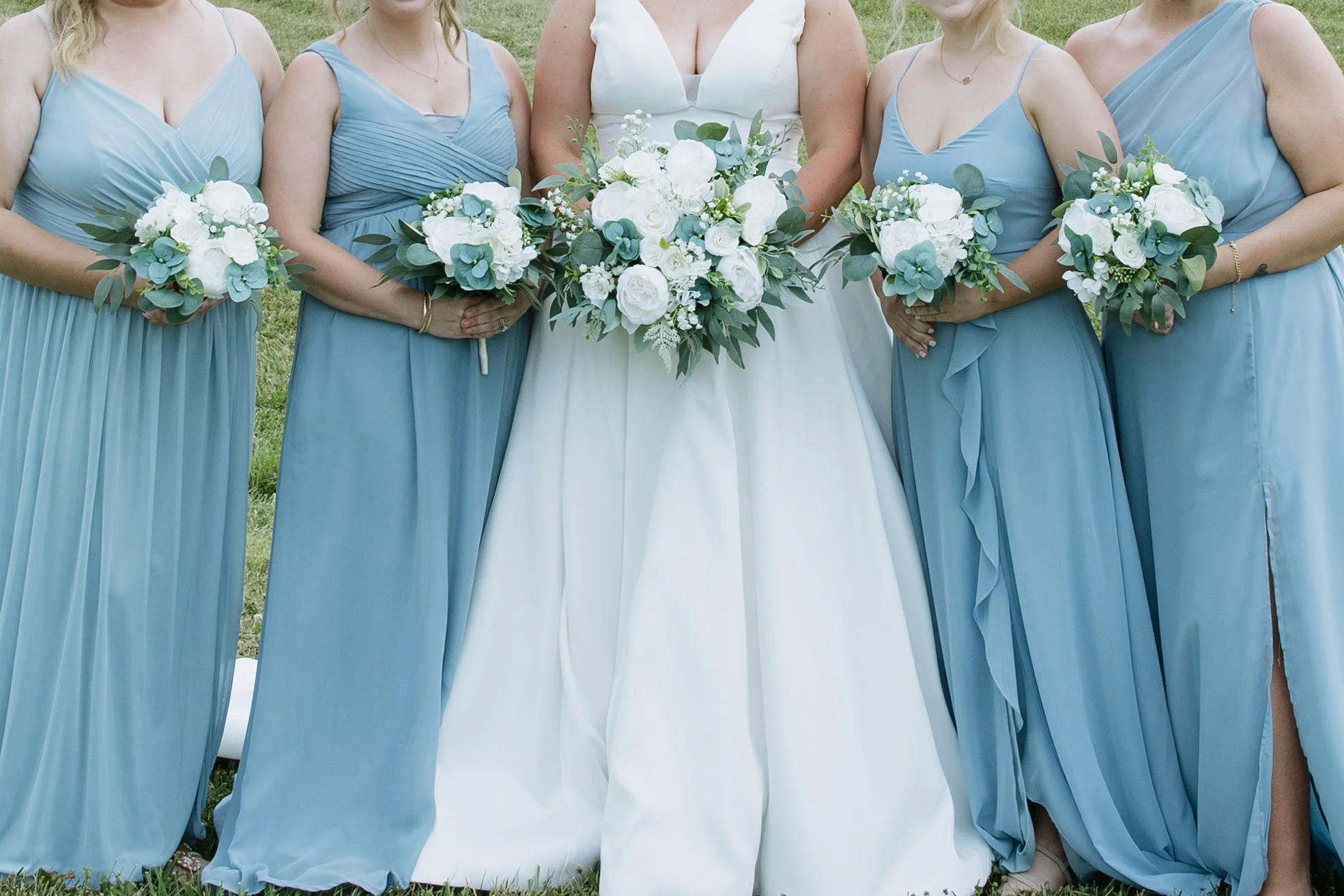 A bride and four bridesmaids standing outdoors, holding bouquets of white flowers and greenery.