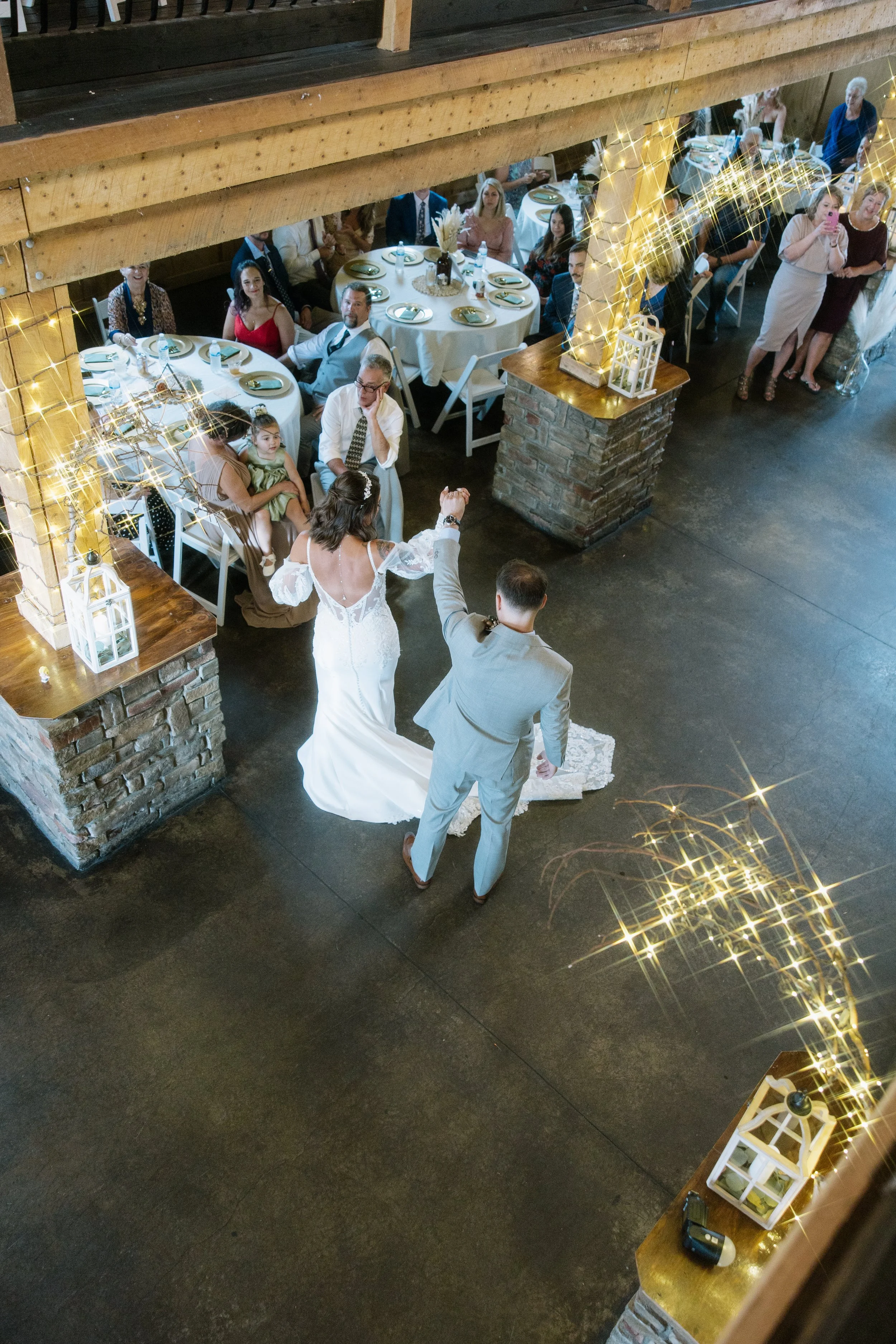 A bride and groom dancing at a wedding reception, with guests seated at decorated tables watching from the background, under string lights and wooden beams.