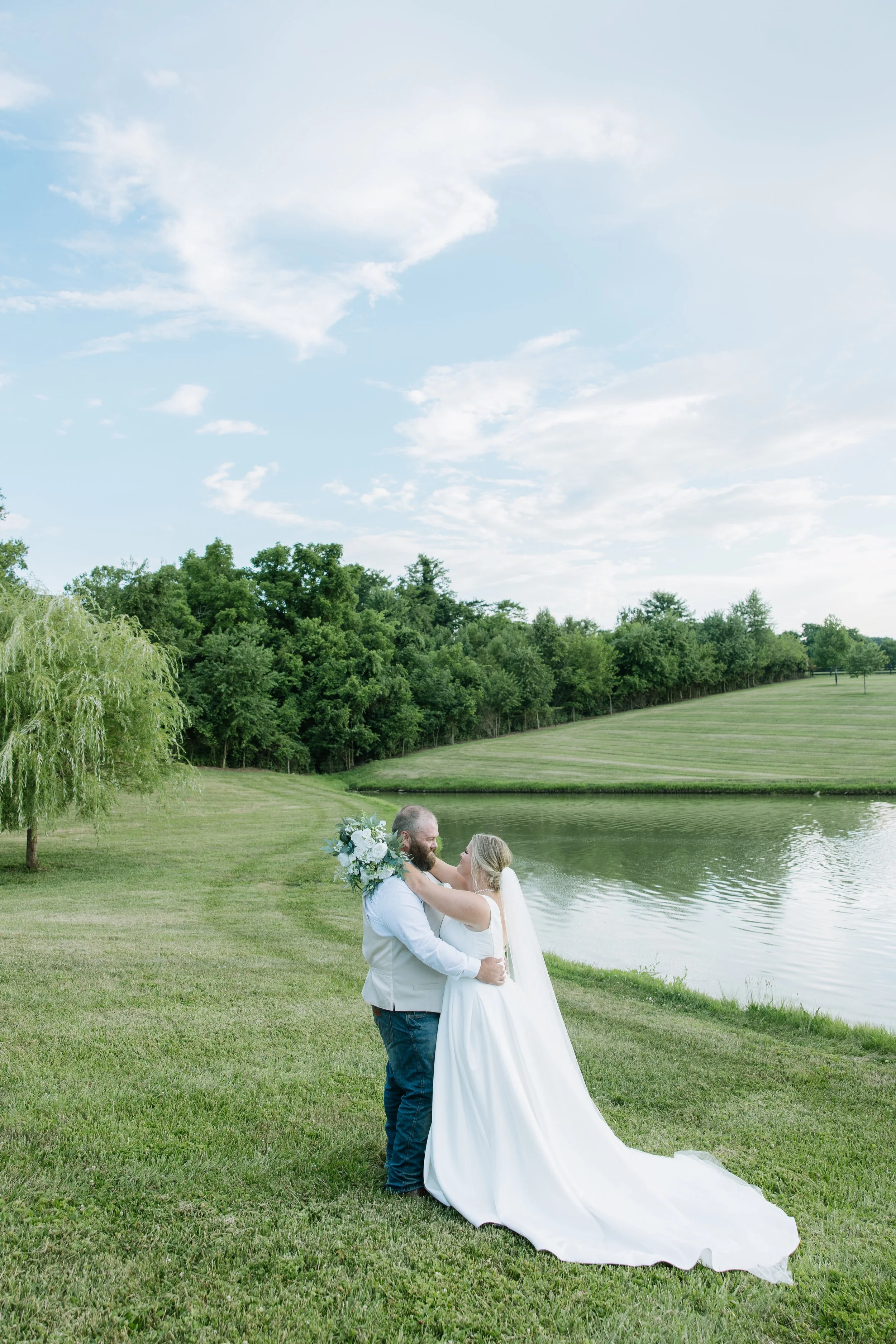 A bride and groom embracing by a lake in a park with green grass, trees, and a partly cloudy sky.