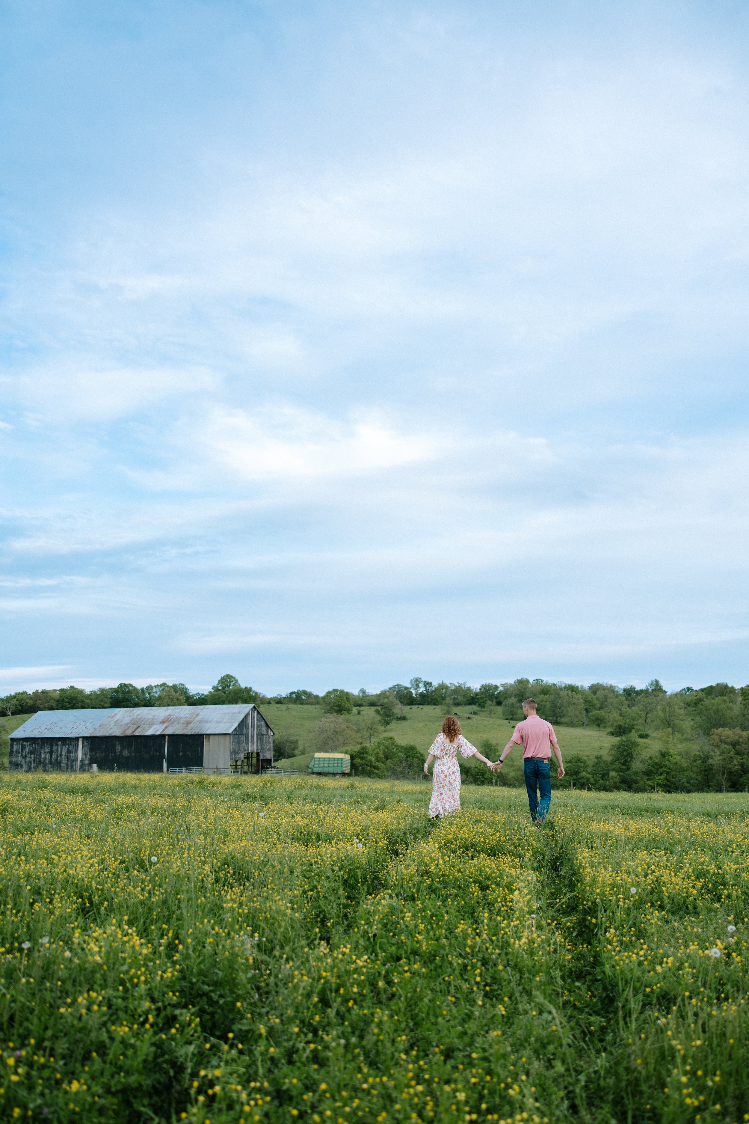 A couple holding hands walking through a yellow flower-covered field towards a rustic barn under a partly cloudy sky.