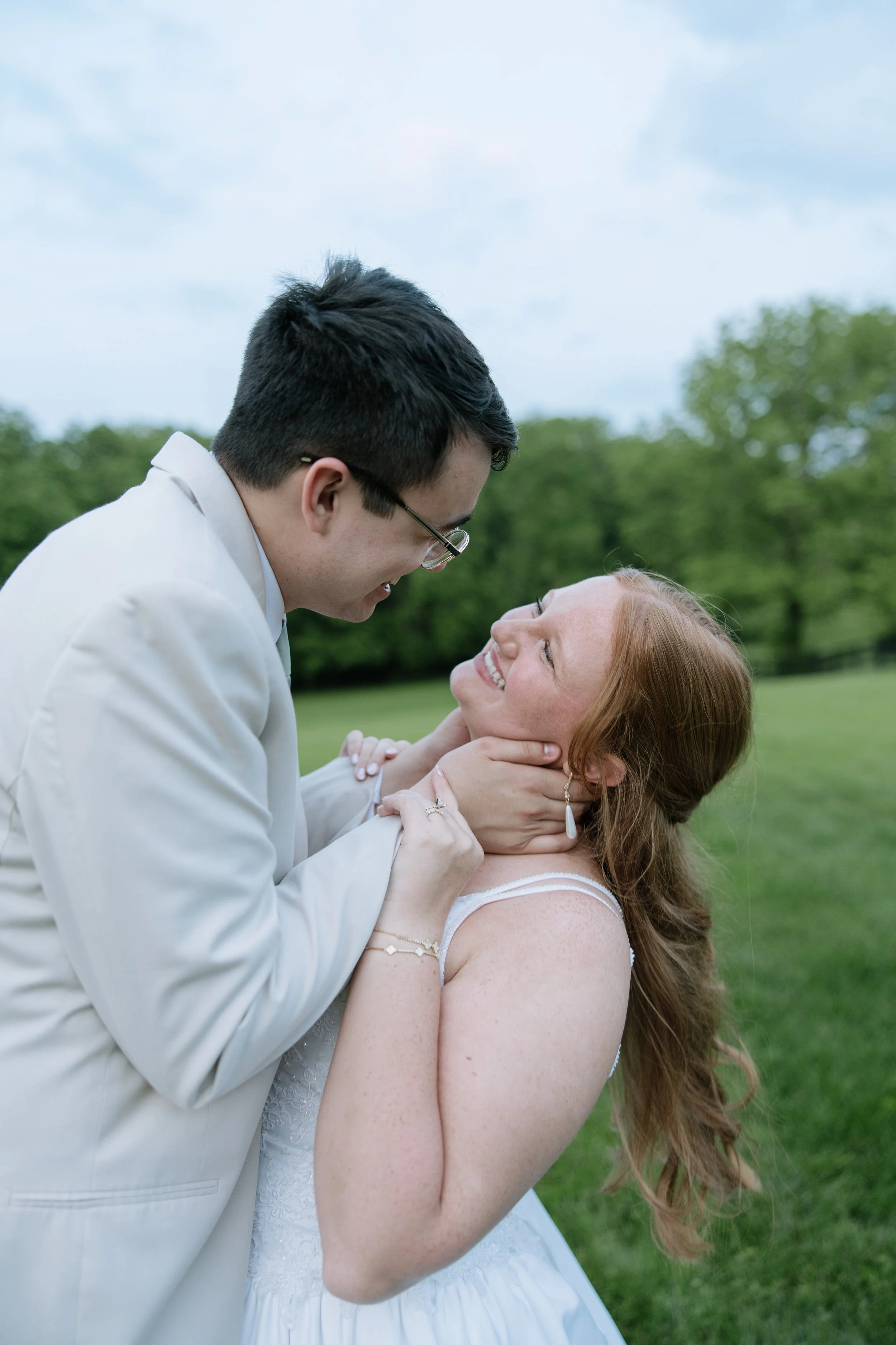 A couple, with the man in a white suit and the woman in a white dress, sharing an intimate moment outdoors on a grassy field with trees in the background.
