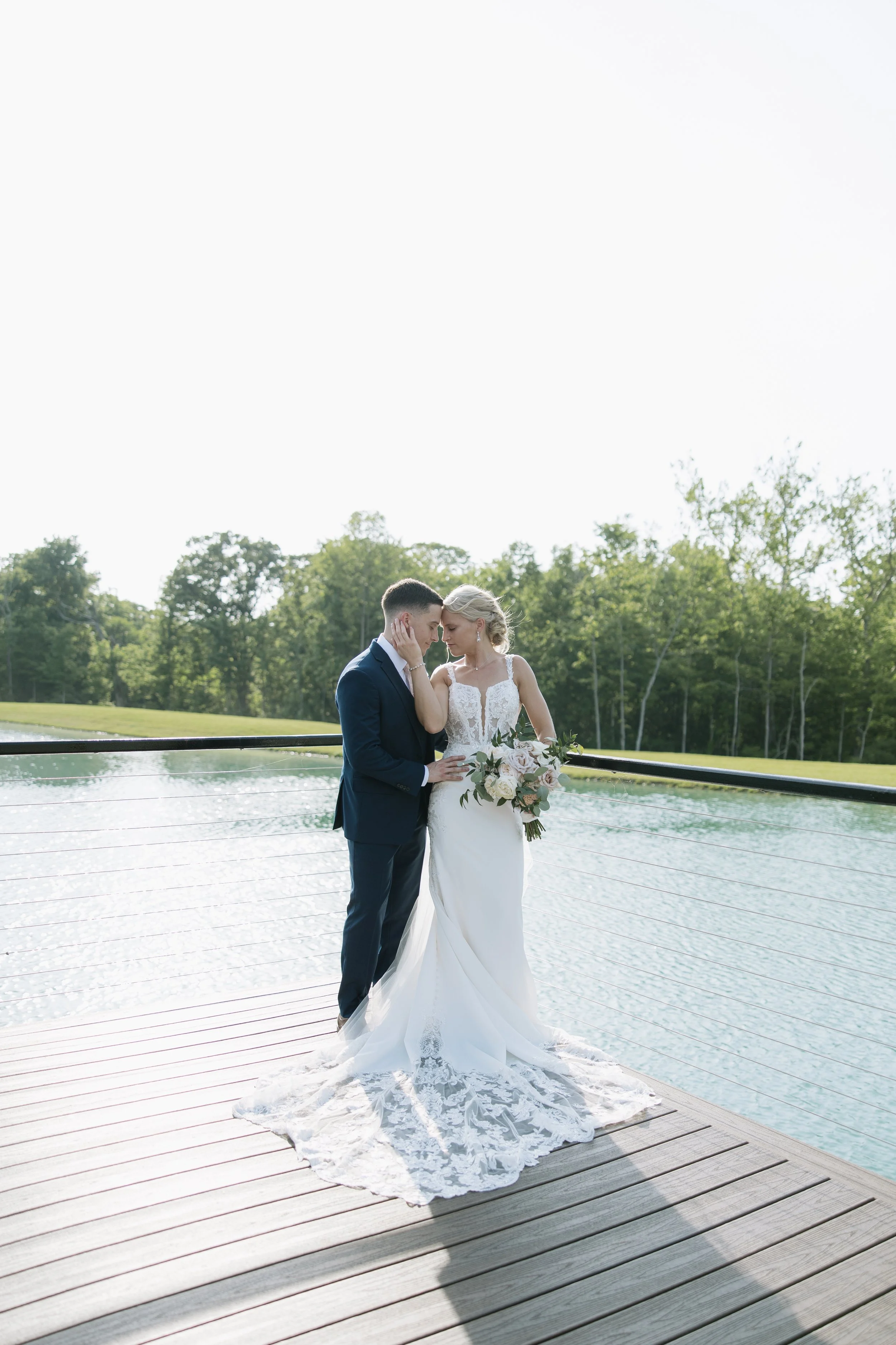 Bride and groom standing close on a dock by a lake, with green trees in the background, during a wedding photoshoot.