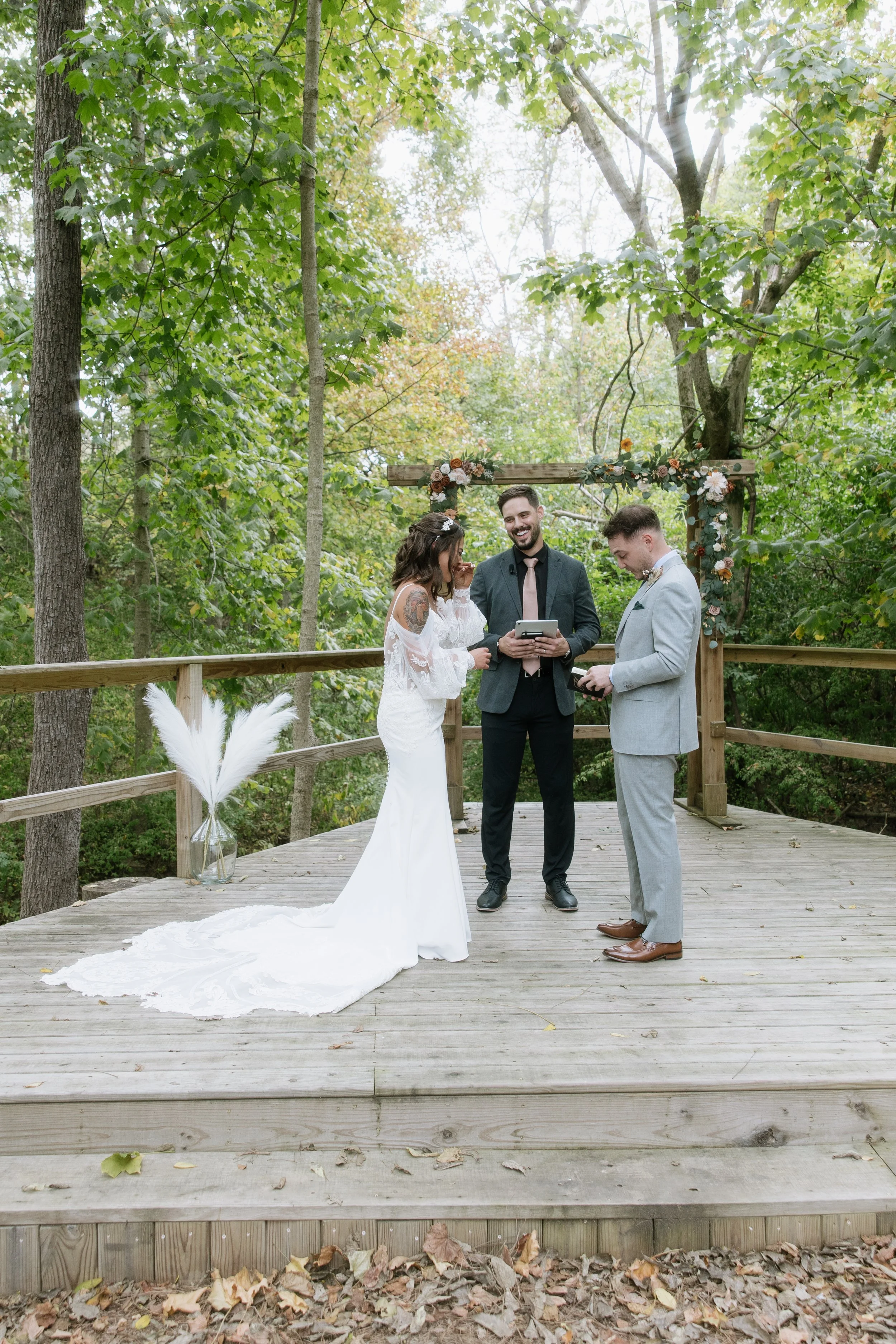 Wedding ceremony taking place outdoors on a wooden platform surrounded by trees, with an officiant, bride, and groom.