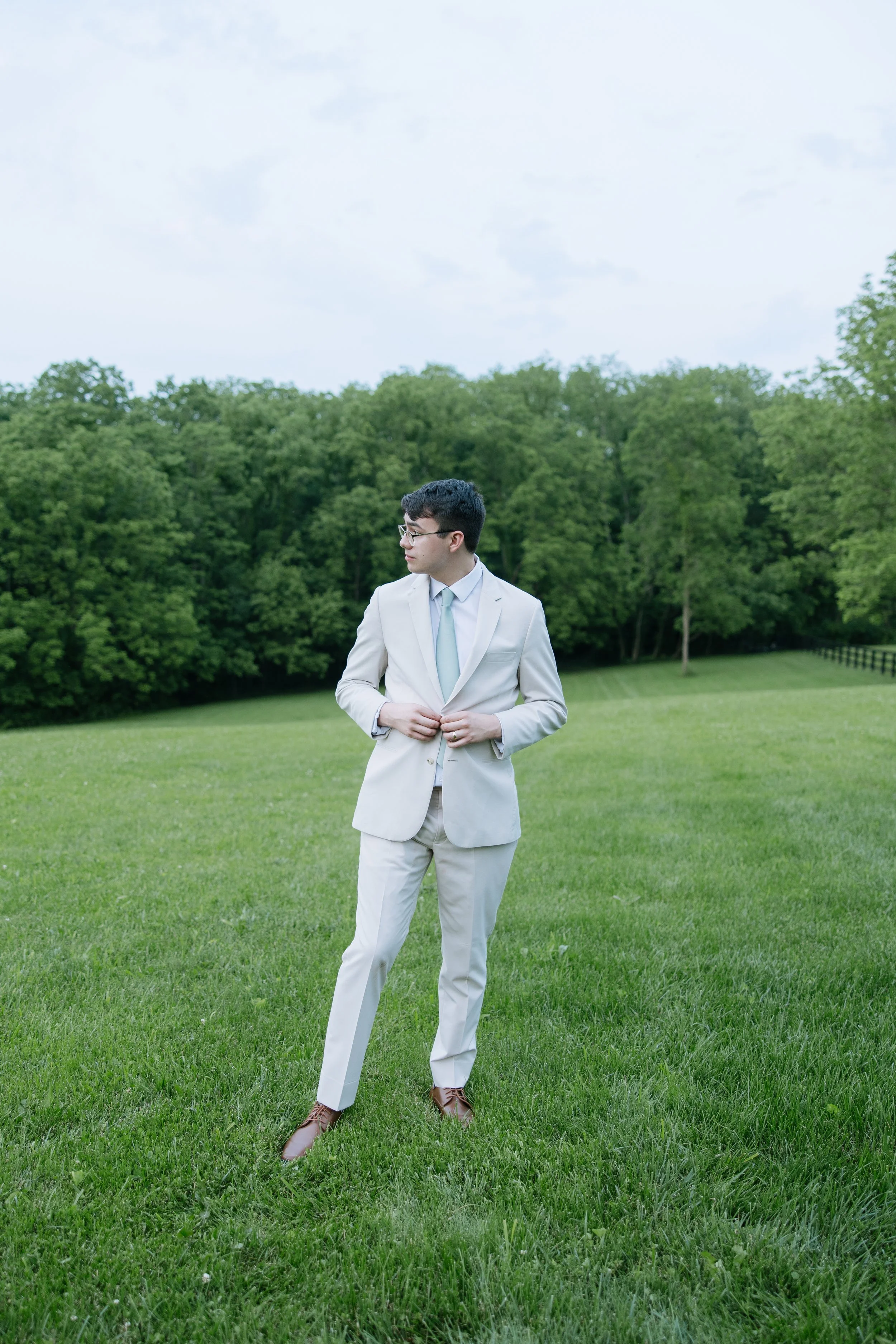 A man wearing a light gray suit, white shirt, and light blue tie stands on a grassy field with trees in the background, adjusting his jacket.