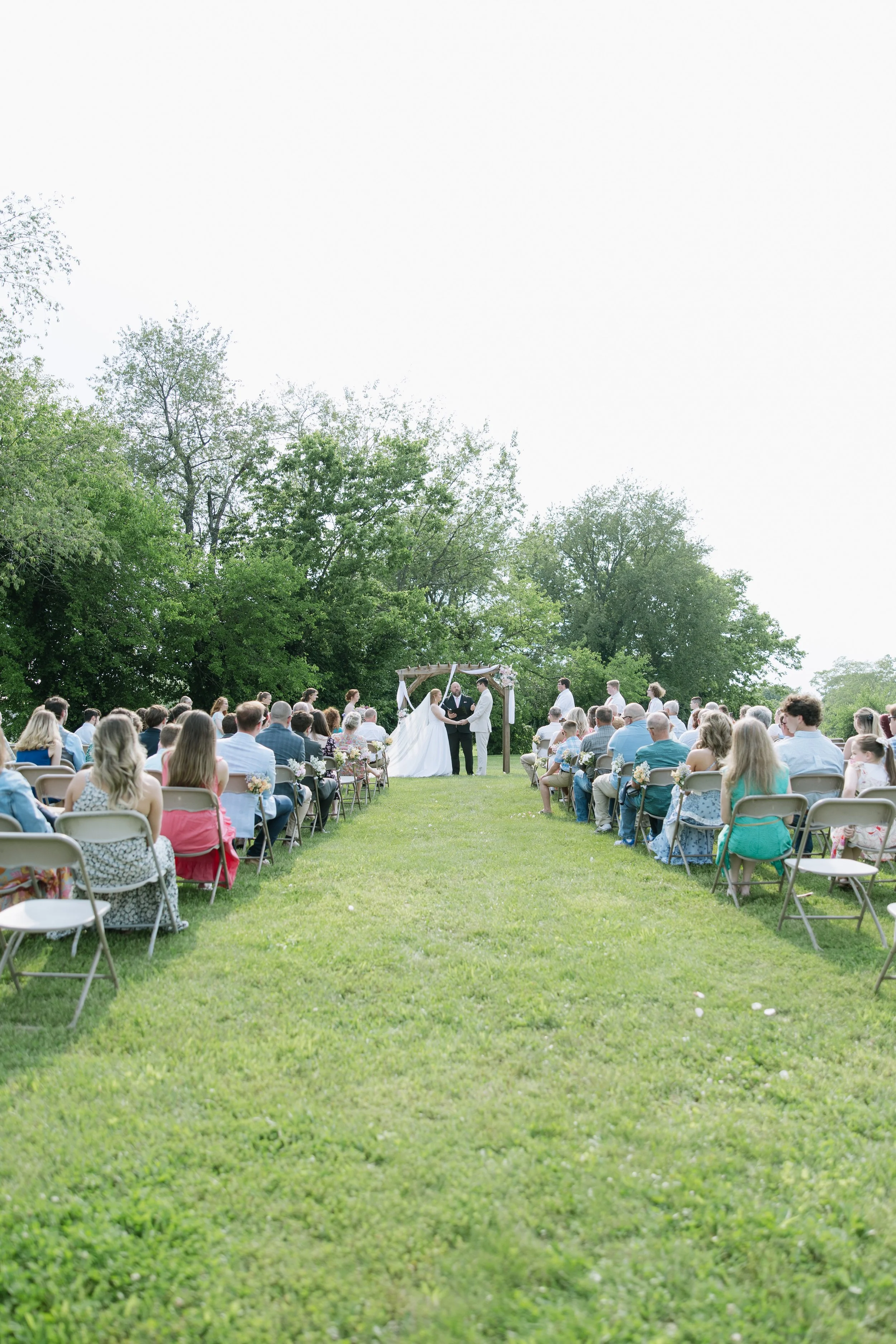 Outdoor wedding ceremony with guests seated on both sides of a grass aisle, couple exchanging vows under a wooden arch, surrounded by trees.