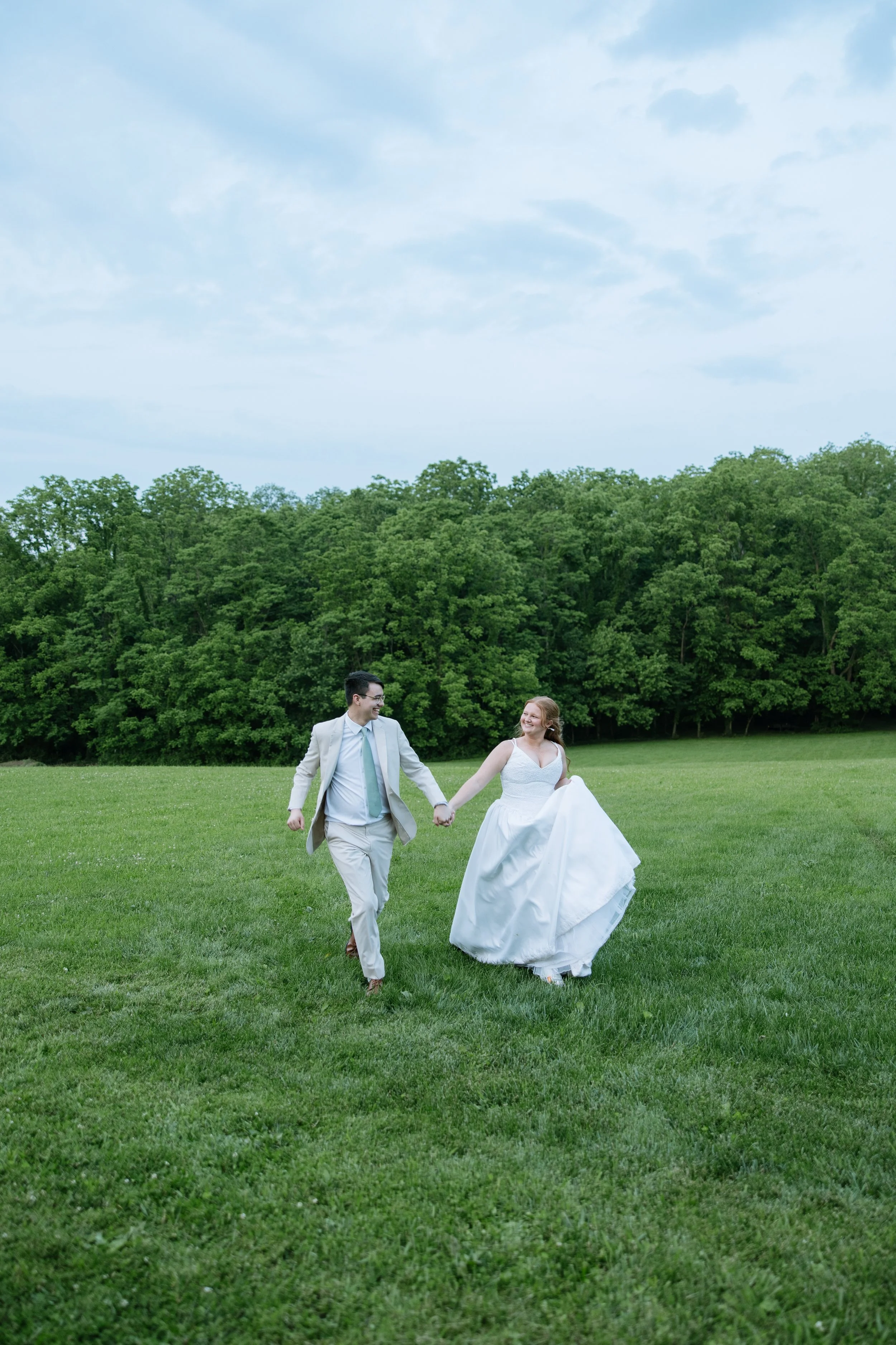 A newlywed couple holding hands and running across a grassy field, with a backdrop of dense green trees and a partly cloudy sky.