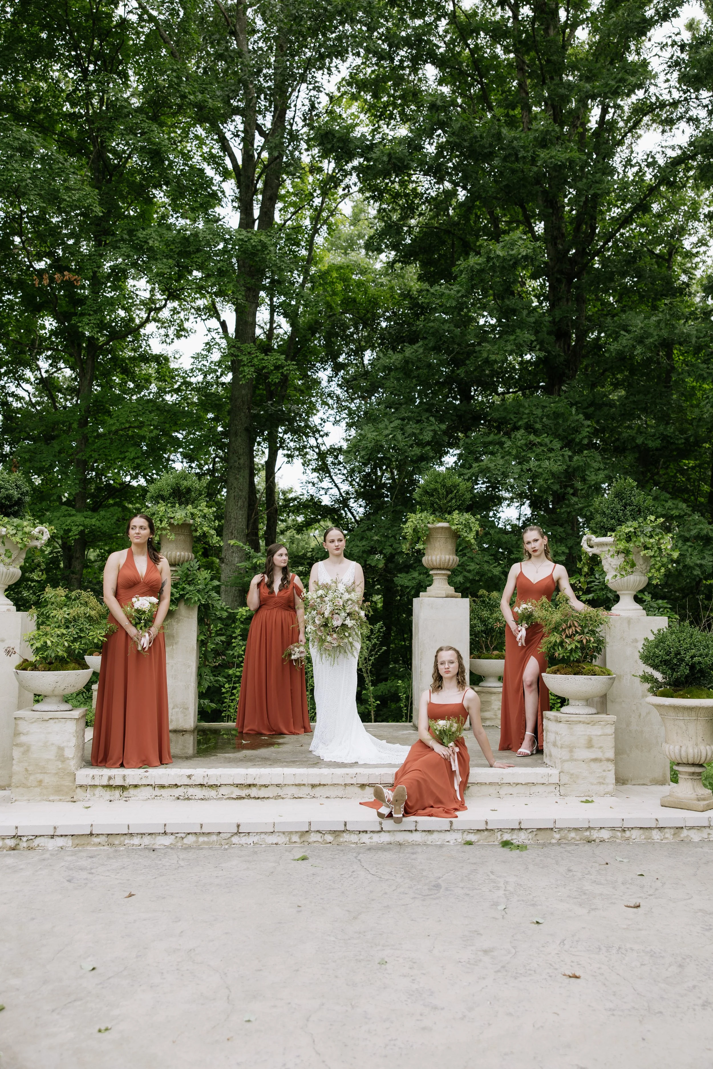 A group of five women in formal dresses, holding bouquets, standing on a stone platform outdoors with lush green trees in the background.