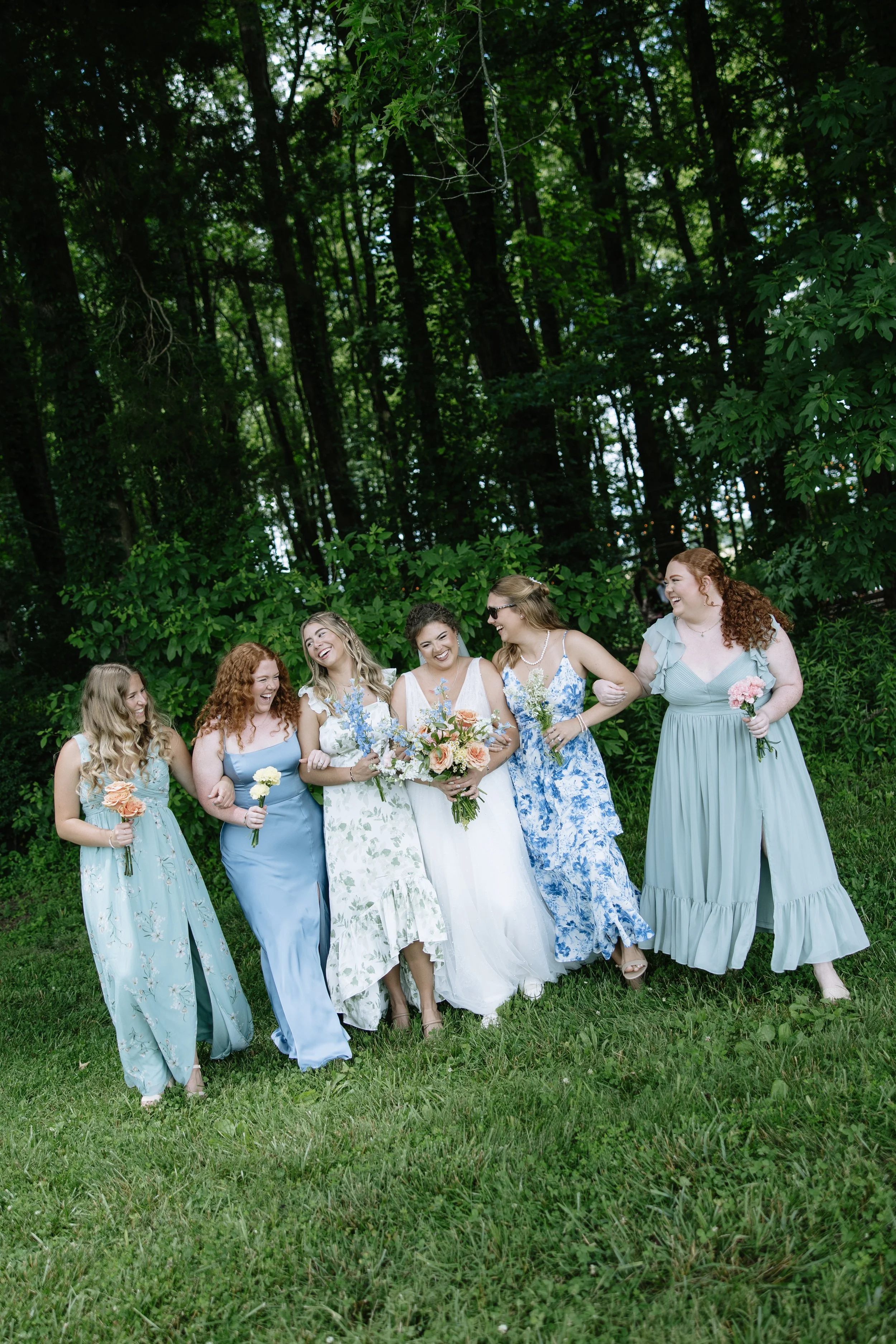 Group of women dressed in pastel-colored dresses, smiling and walking together outdoors in a green, wooded area, holding flowers.