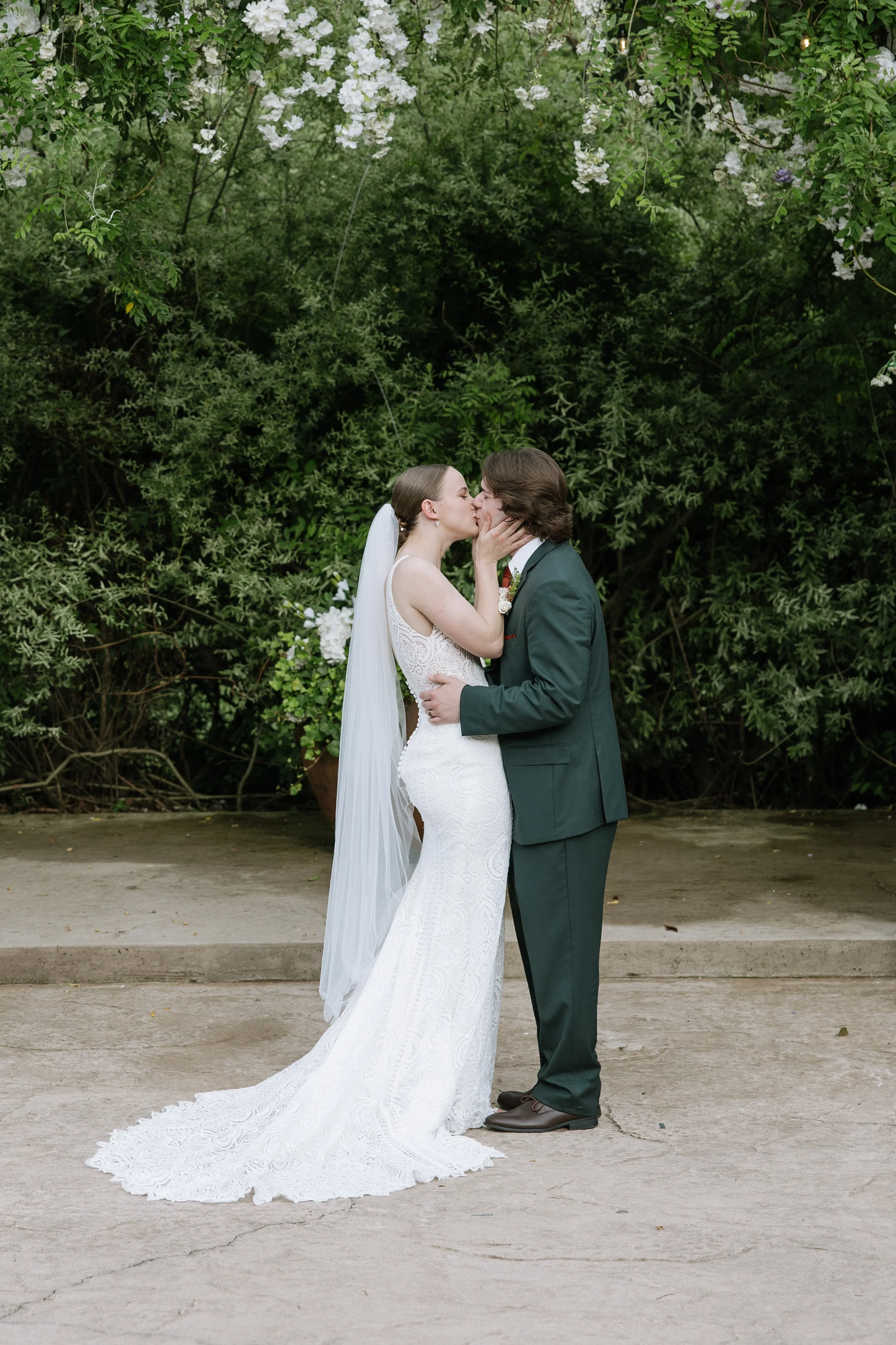 A bride and groom kissing outdoors, surrounded by green foliage and flowering plants, with the bride wearing a white lace wedding gown and veil, and the groom in a dark green suit.