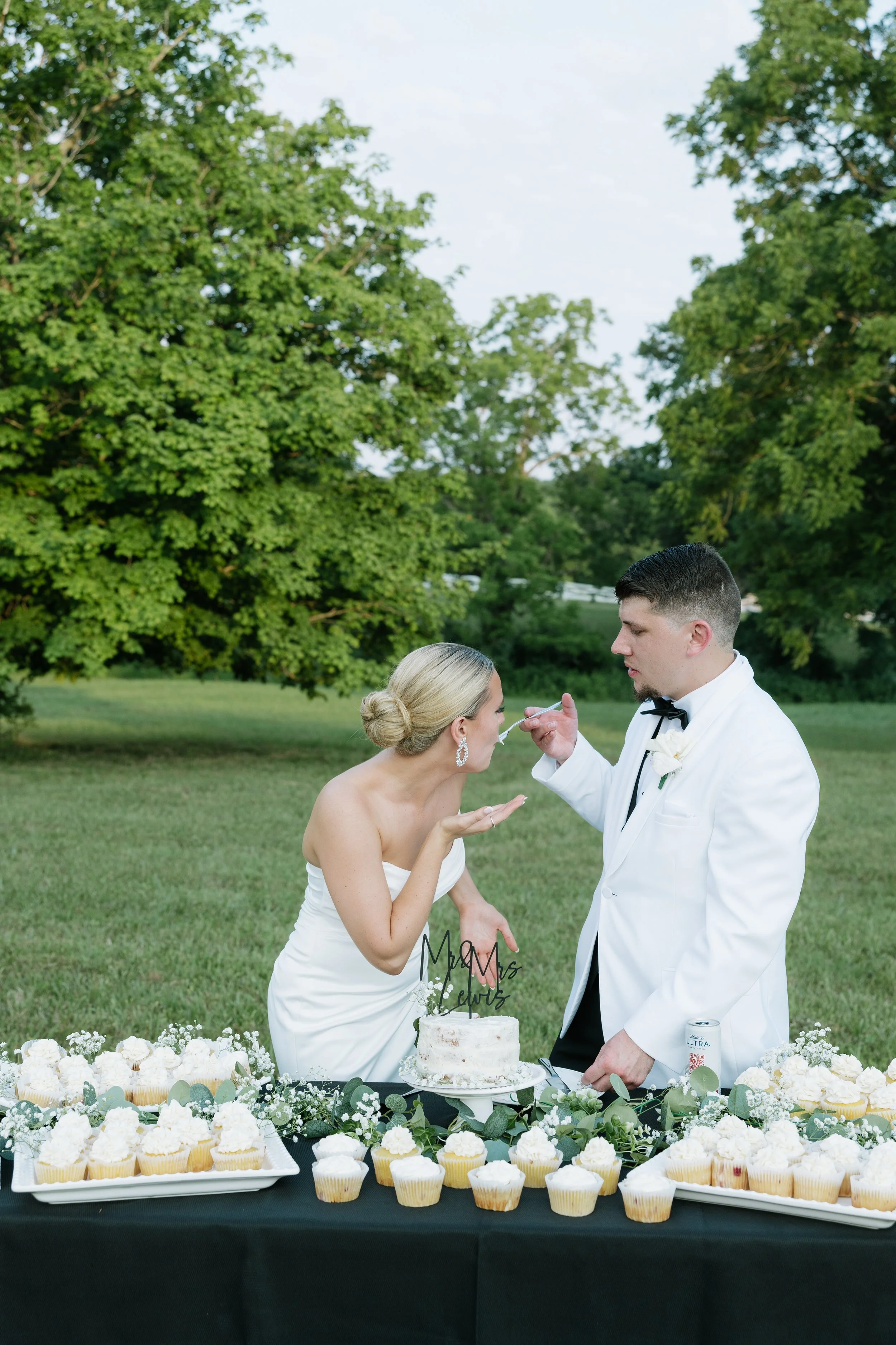 Bride and groom at their outdoor wedding reception, with the bride blowing out candles on a small wedding cake while the groom helps, surrounded by cupcakes and greenery.