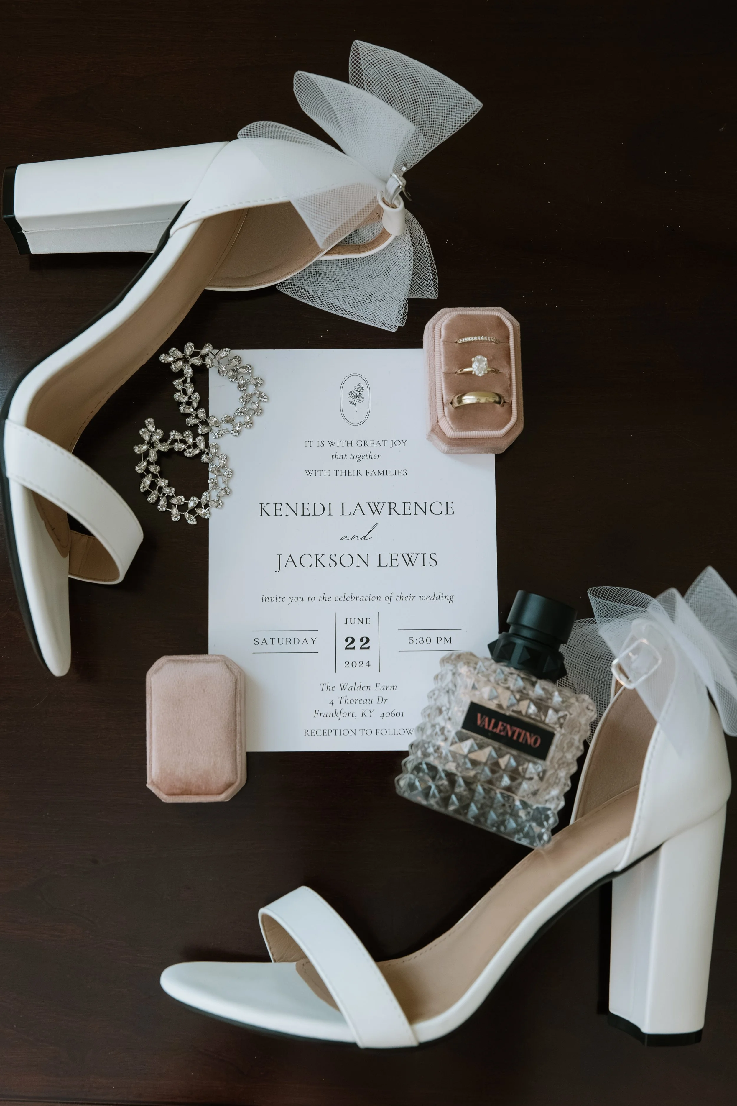 Wedding invitation surrounded by white high-heeled shoes, pearl necklace, ring box, perfume, and jewelry on a dark wooden surface.