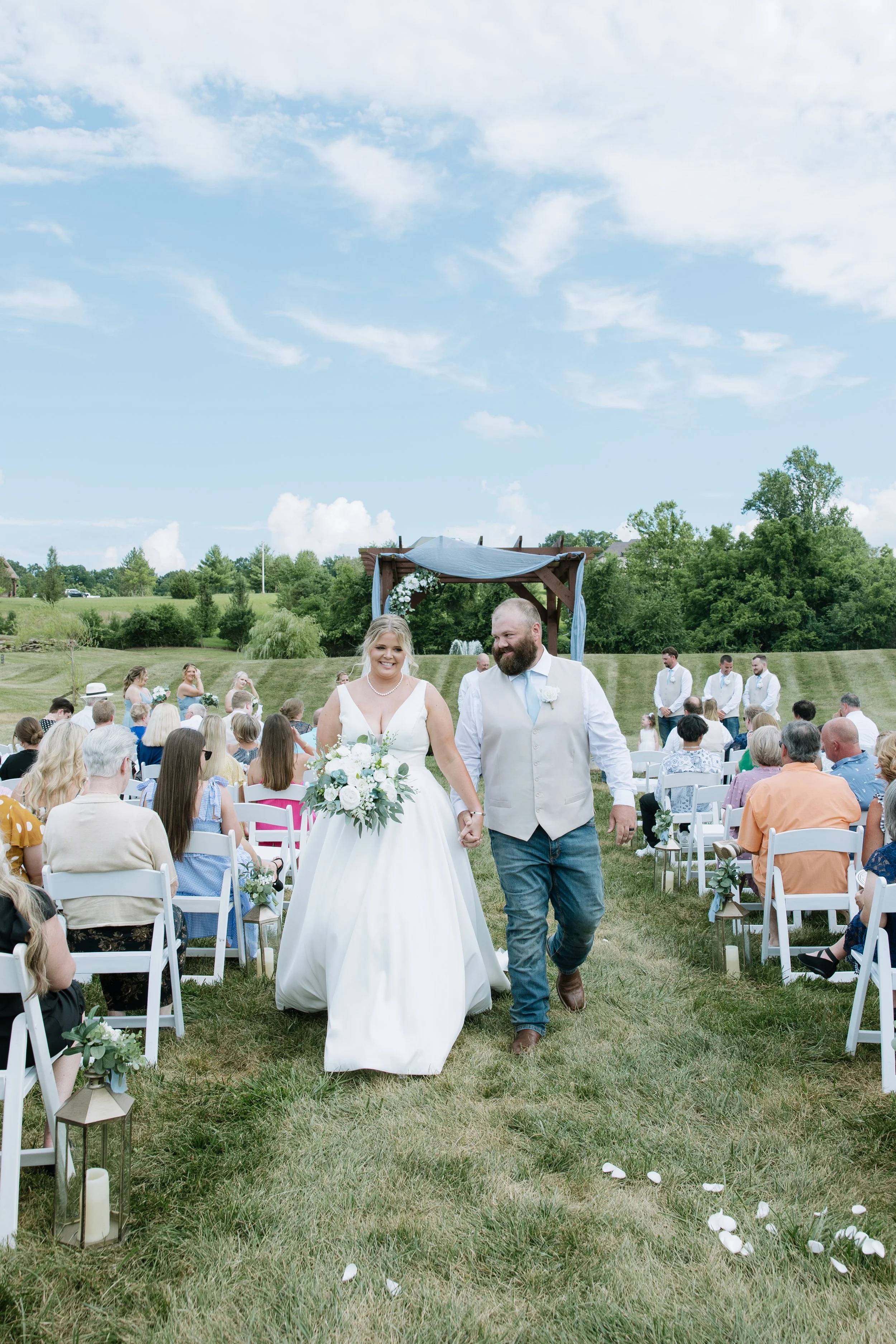 A bride and groom walking hand in hand down the aisle at their outdoor wedding ceremony surrounded by seated guests, with a decorative arch in the background and a bright, partly cloudy sky.