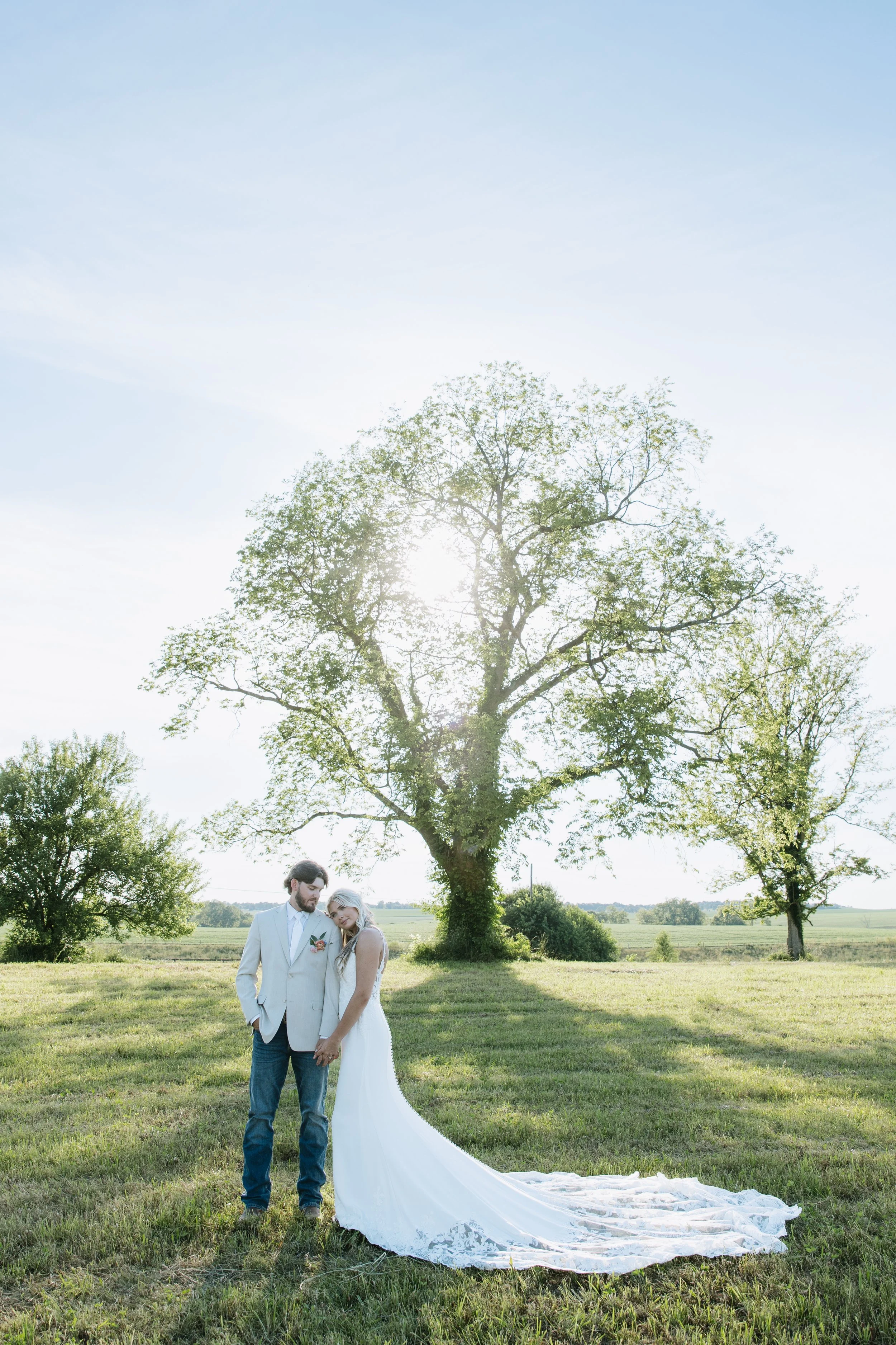 A wedding couple standing outdoors in a grassy field with a large tree and clear sky behind them, sunlight shining through the leaves, the bride wearing a white wedding gown with a long train, and the groom in a white blazer and jeans.
