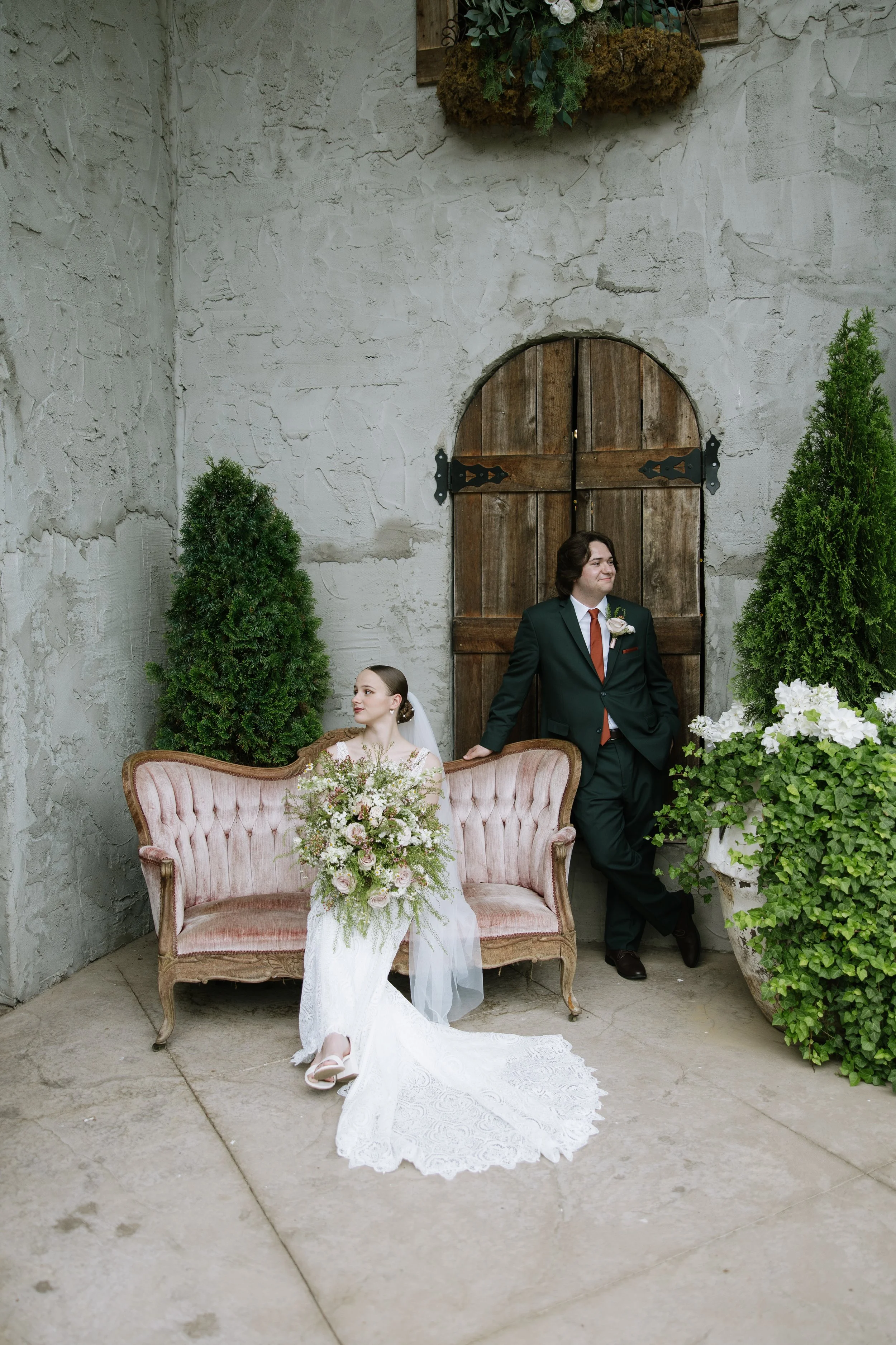 A bride in a white wedding dress and veil sitting on a vintage pink sofa holding a bouquet, and a groom in a dark suit with a white flower boutonniere leaning against a wooden door with a split flourish. They are surrounded by greenery and white flow