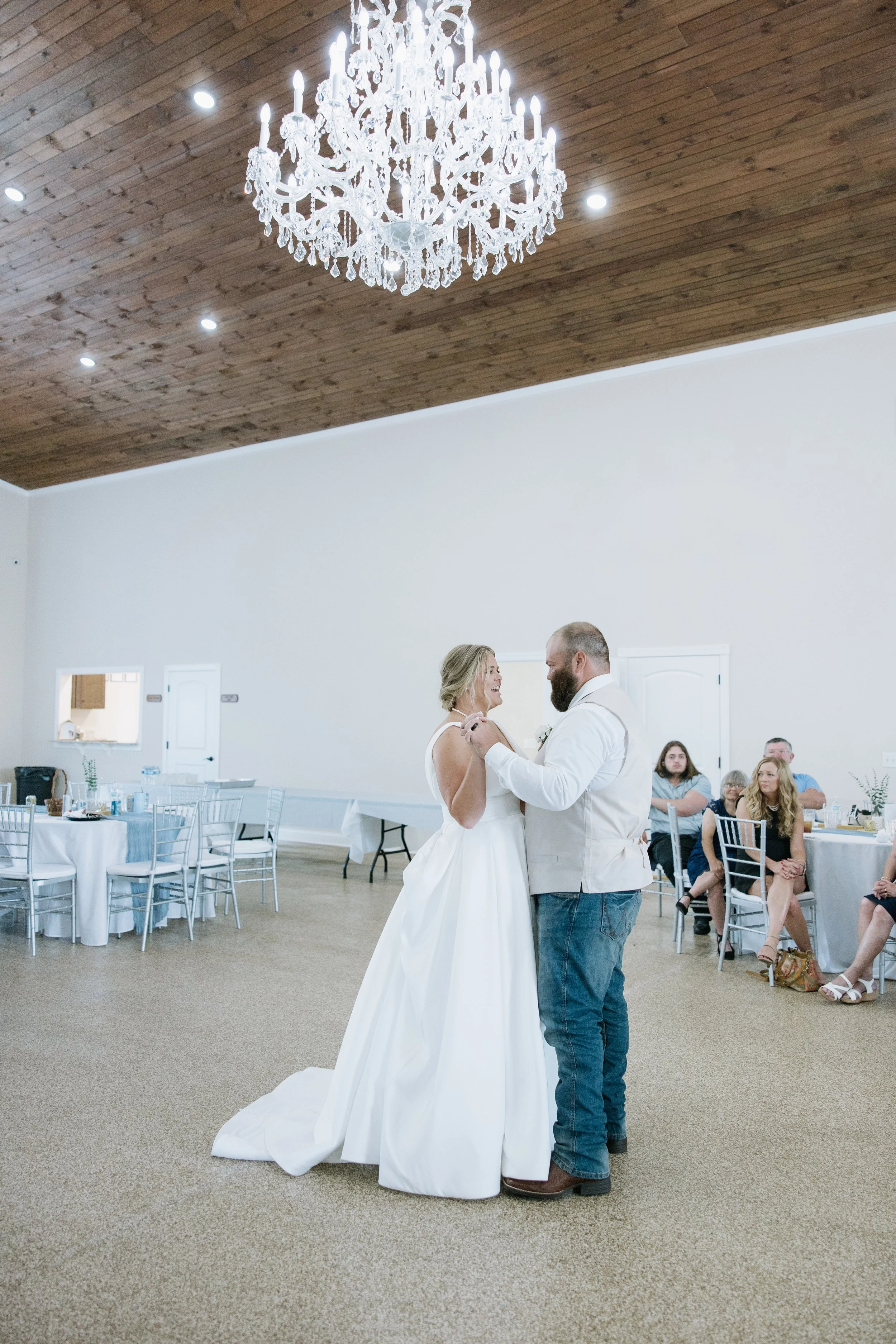 A bride and groom dance during their wedding reception in an elegant hall with a high wooden ceiling and a large chandelier, with guests seated at round tables in the background.