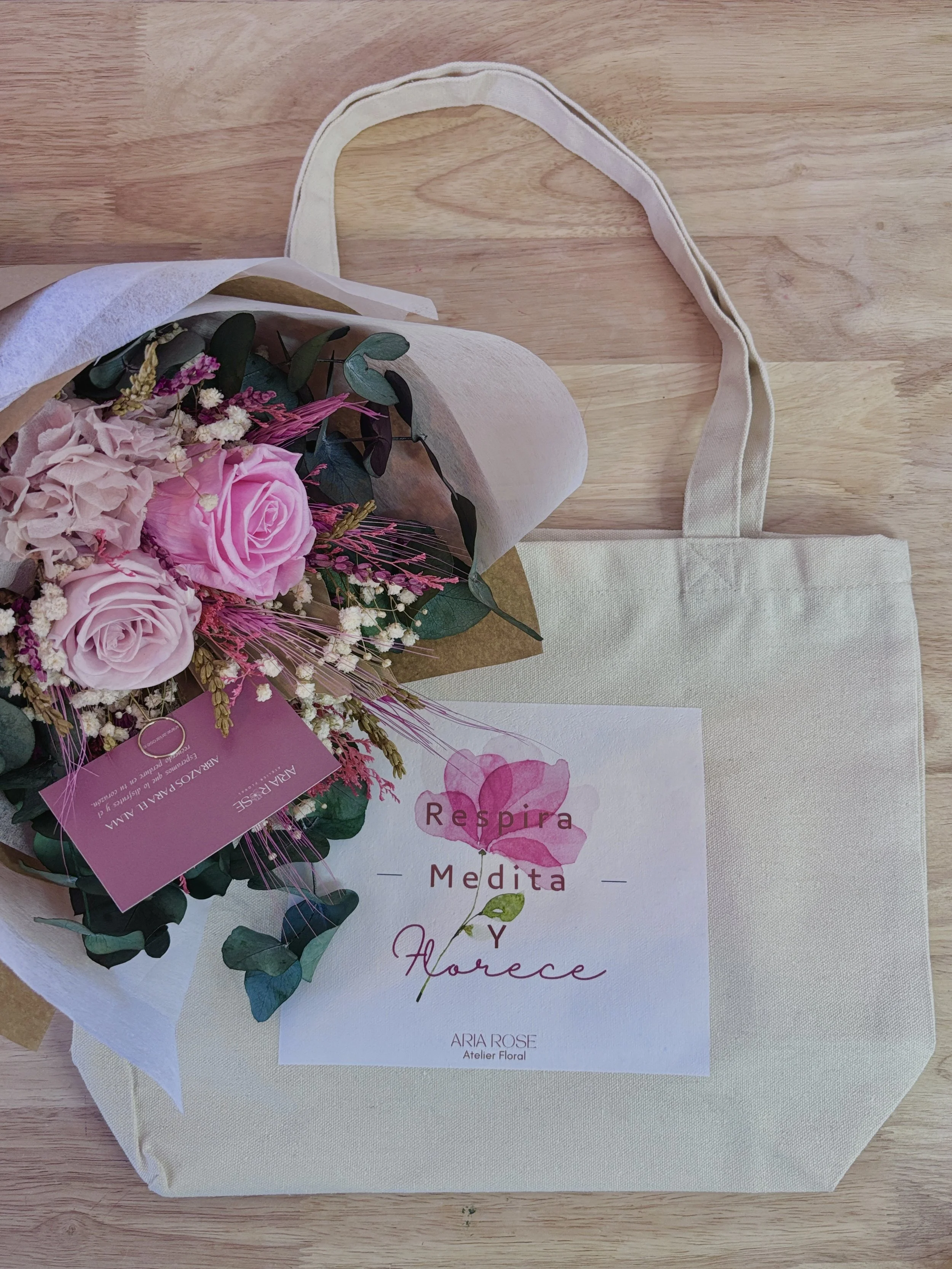 Un ramo de flores con rosas rosas y otras flores pequeñas, acompañadas de una bolsa de tela beige con un cartel que dice 'Respira, Medita y Florece' de ARIA ROSE, Atelier Floral.