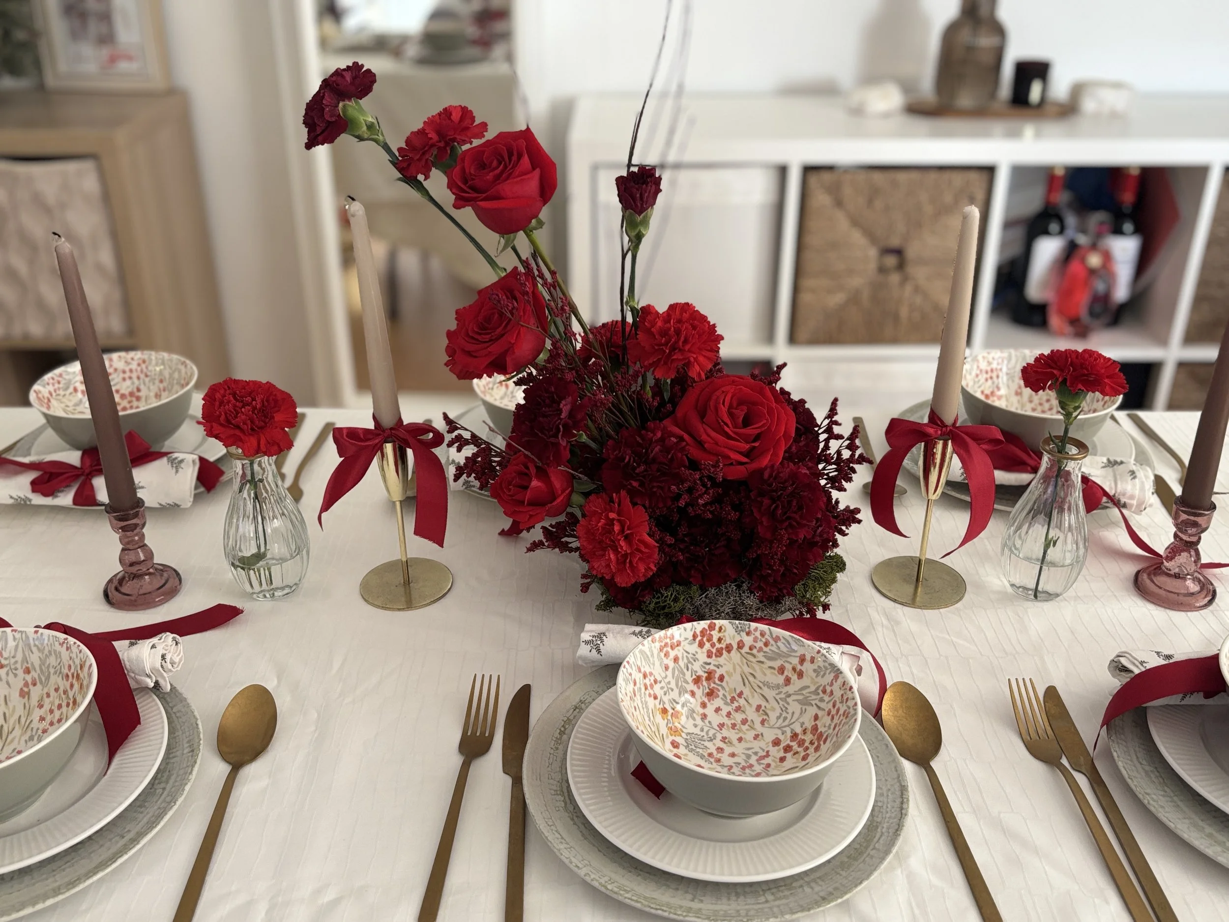 Mesa decorada con flores rojas, platos, cubiertos dorados, velas en candelabros y mantelería blanca.
