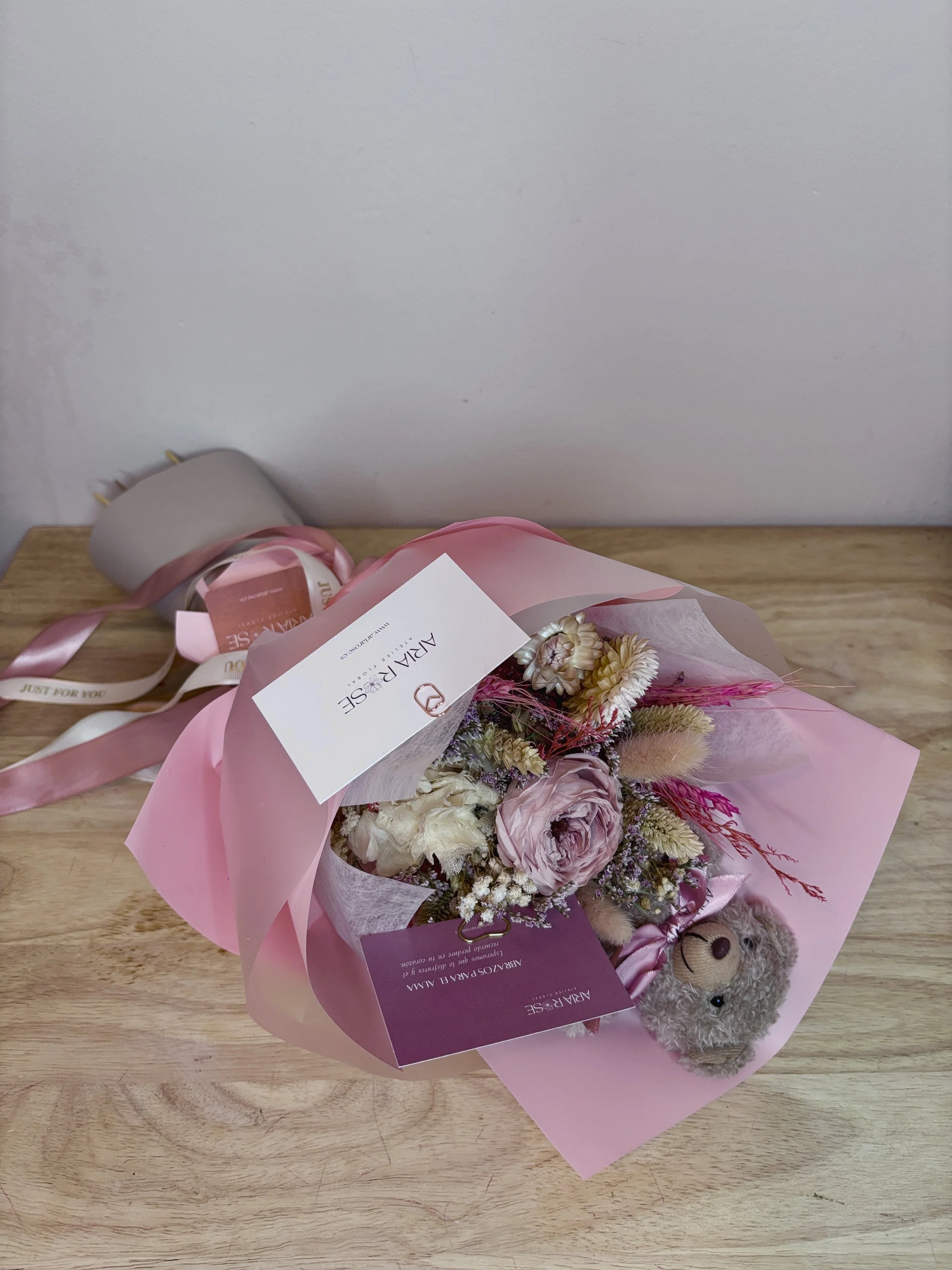 Un ramo de flores en tonos rosa y blanco con un peluche de oso, decorado con cintas rosas, sobre una mesa de madera.