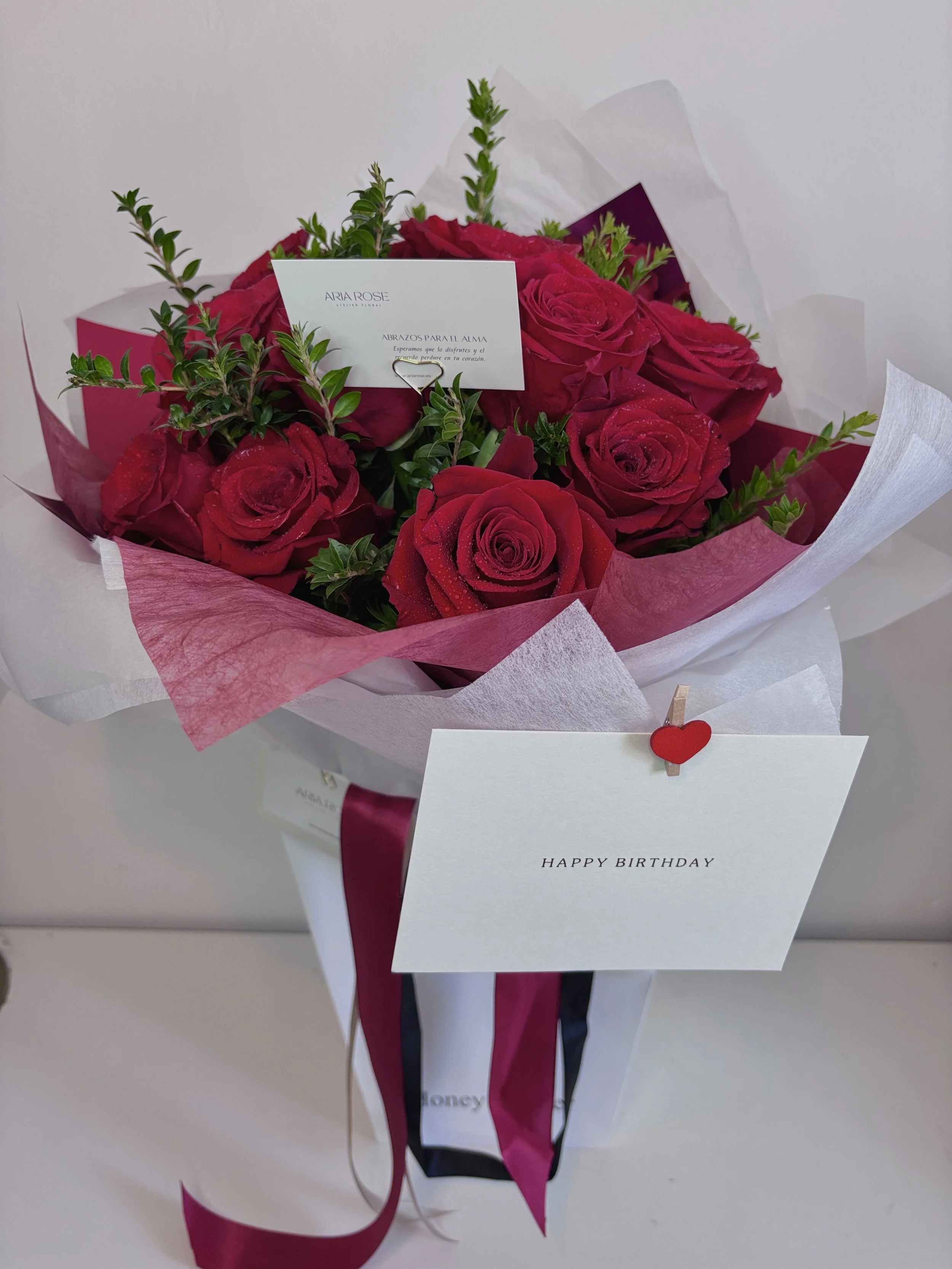 Un ramo de rosas rojas decorado con hojas verdes, envoltorios blancos, rosa y morado, acompañado de una tarjeta que dice 'HAPPY BIRTHDAY' y otra tarjeta con mensaje en español. Hay cintas rosadas, blancas y negras decorando el ramo.