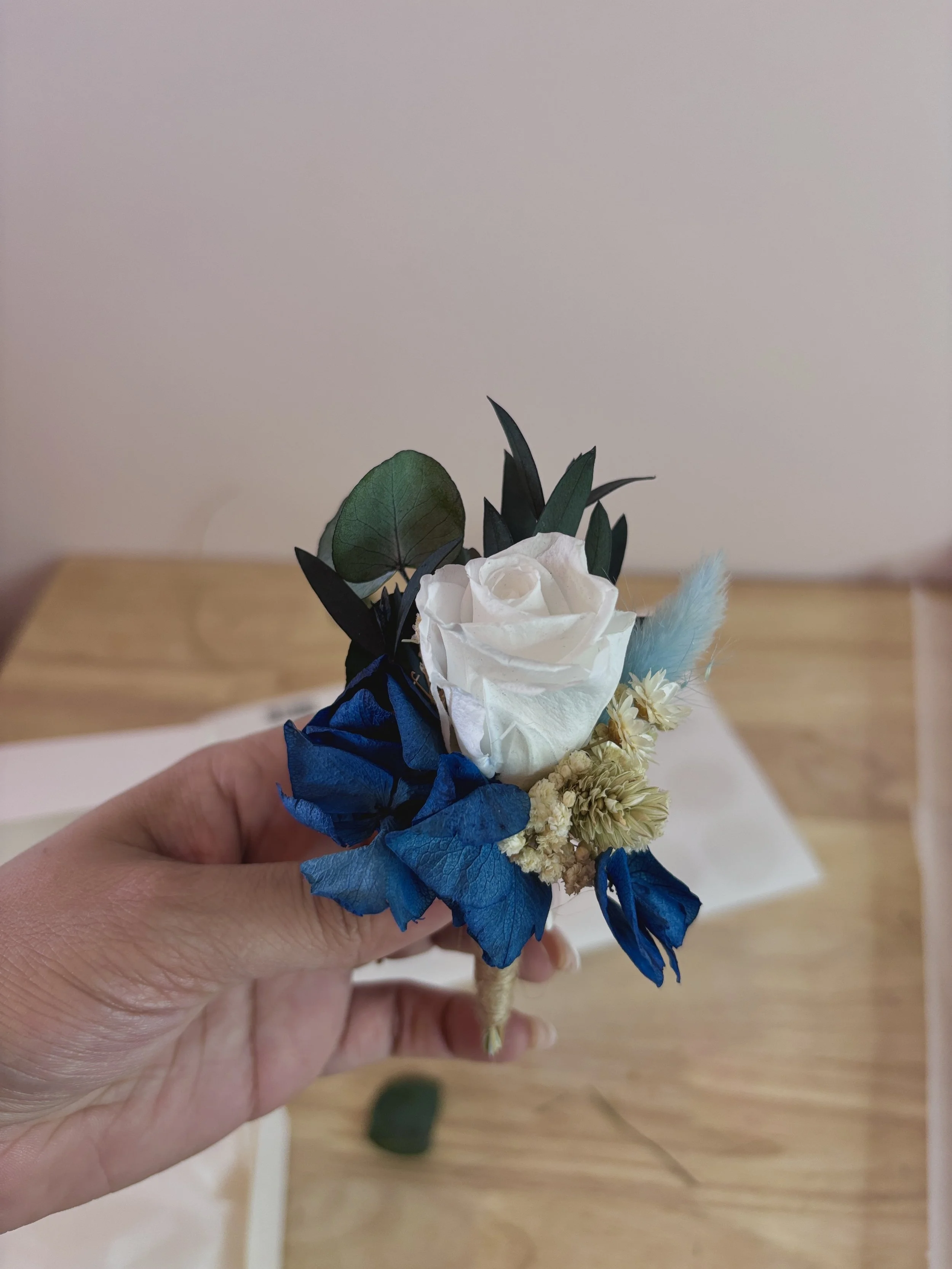 Mano sosteniendo un pequeño arreglo floral con rosa blanca, hojas verdes, flores secas y accesorios azules.