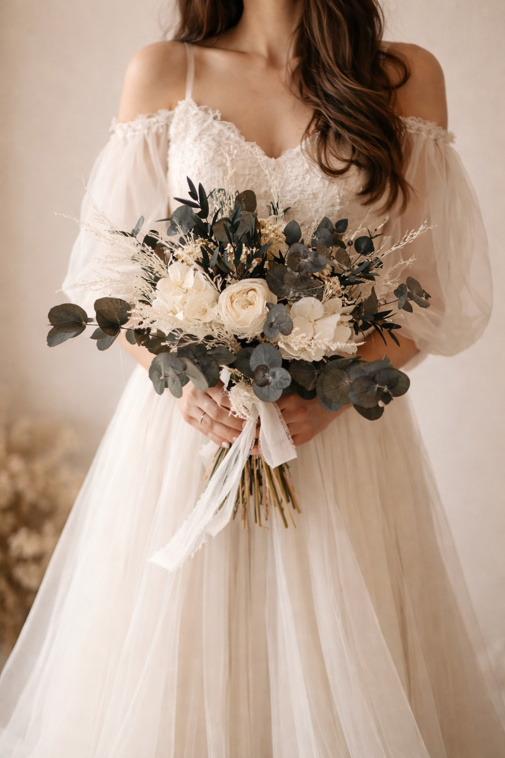 Mujer con vestido de novia blanco, sosteniendo un ramo de flores en tonos blancos, con hojas verdes y follaje seco, en un fondo neutro.