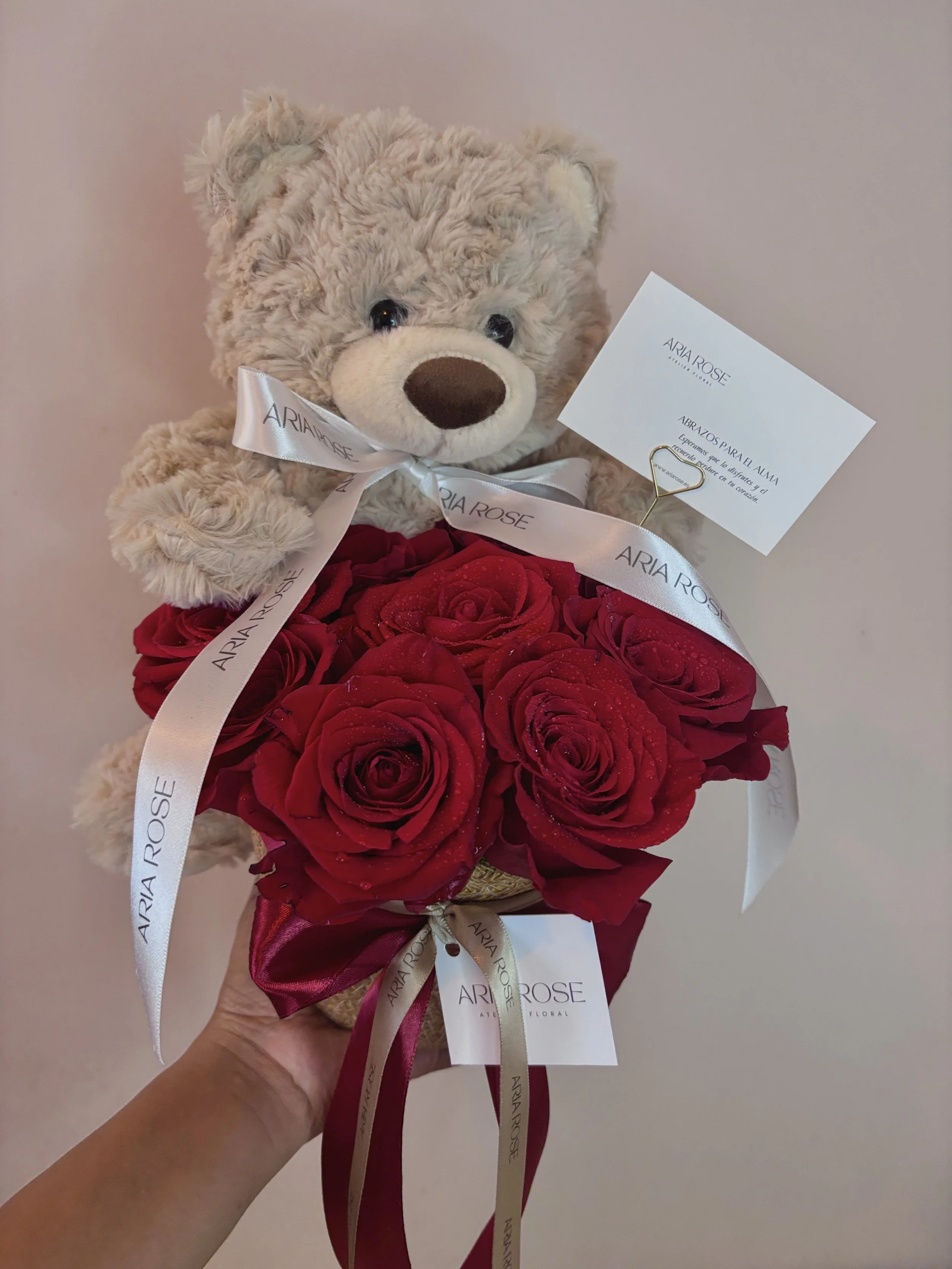 Un ramo de rosas rojas con un oso de peluche sentado sobre ellas, con un lazo blanco que dice 'ARIA ROSE' y una tarjeta adjunta.