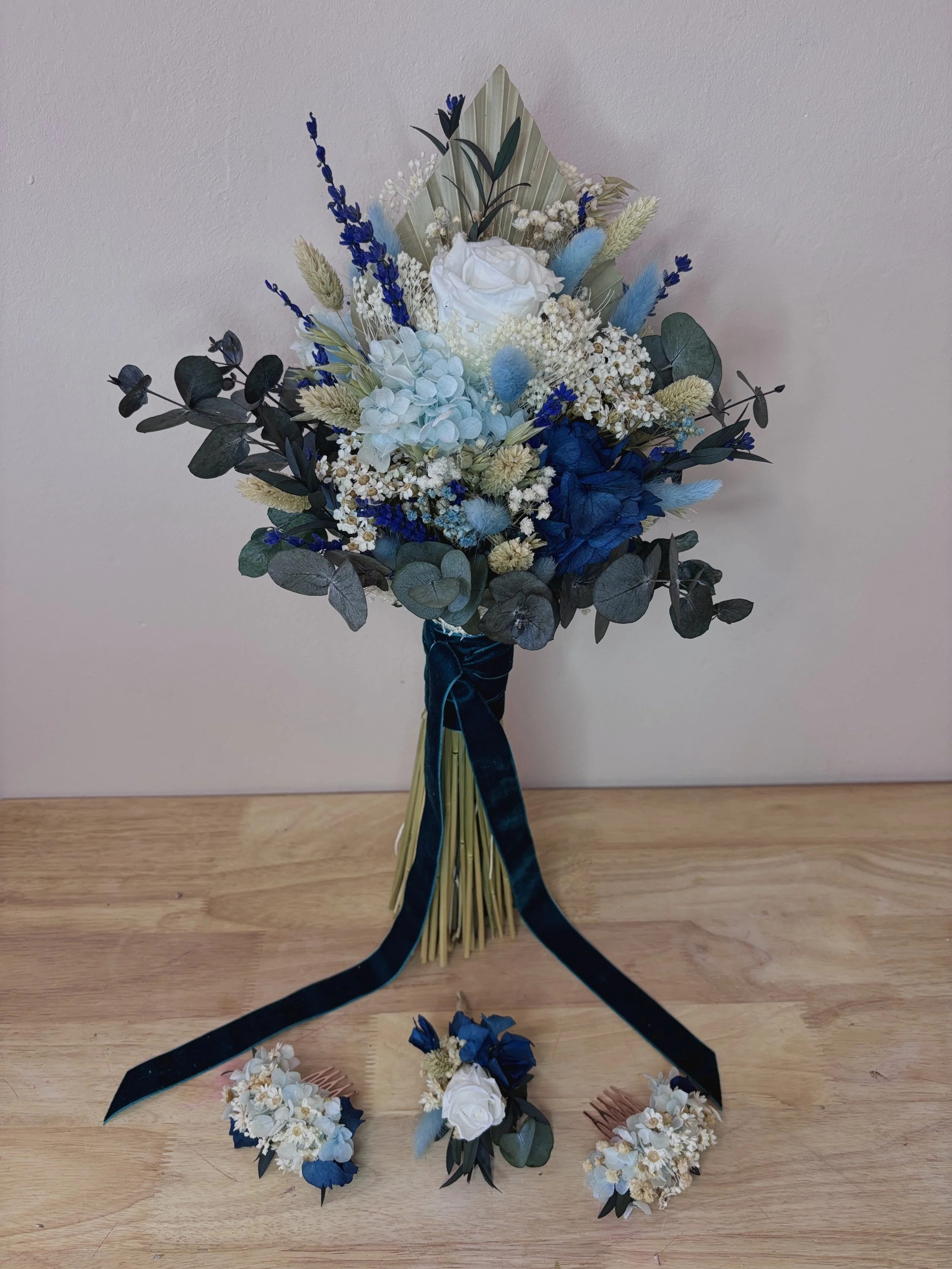 Ramo de flores con rosas blancas y azuladas, eucalipto, lavanda, y otros arreglos florales en tonos blancos y azules, decorado con cinta negra, acompañado de dos pequeños arreglos florales.