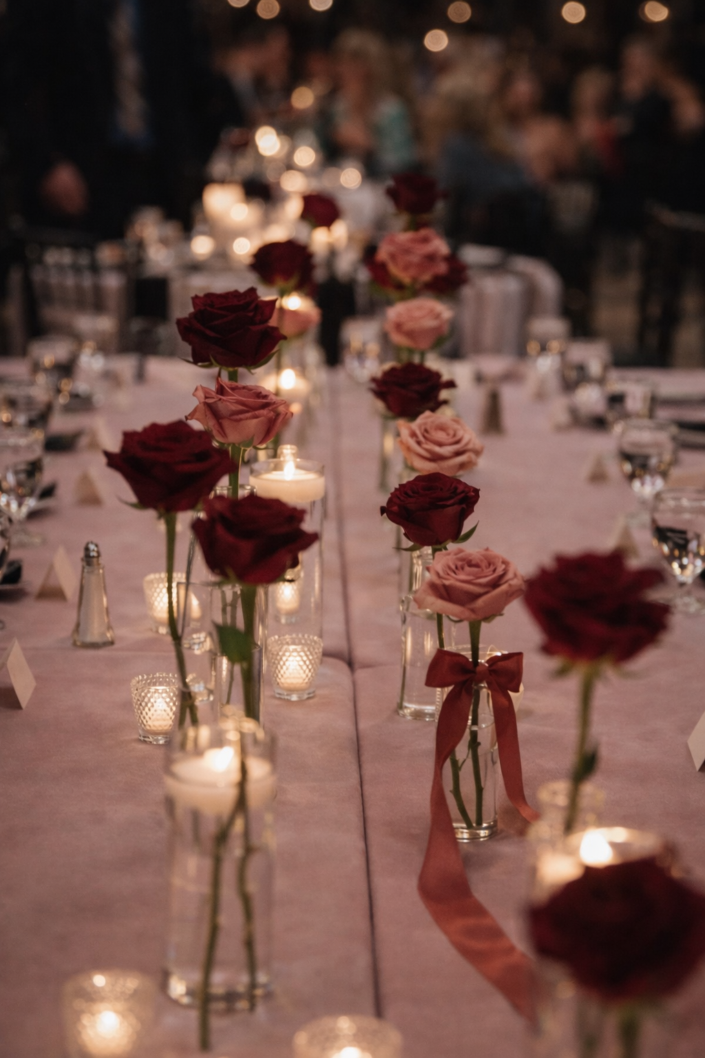 Mesa decorada con rosas rojas y rosas en jarrones, con velas y centros de mesa iluminados, en un ambiente de celebración o evento nocturno.