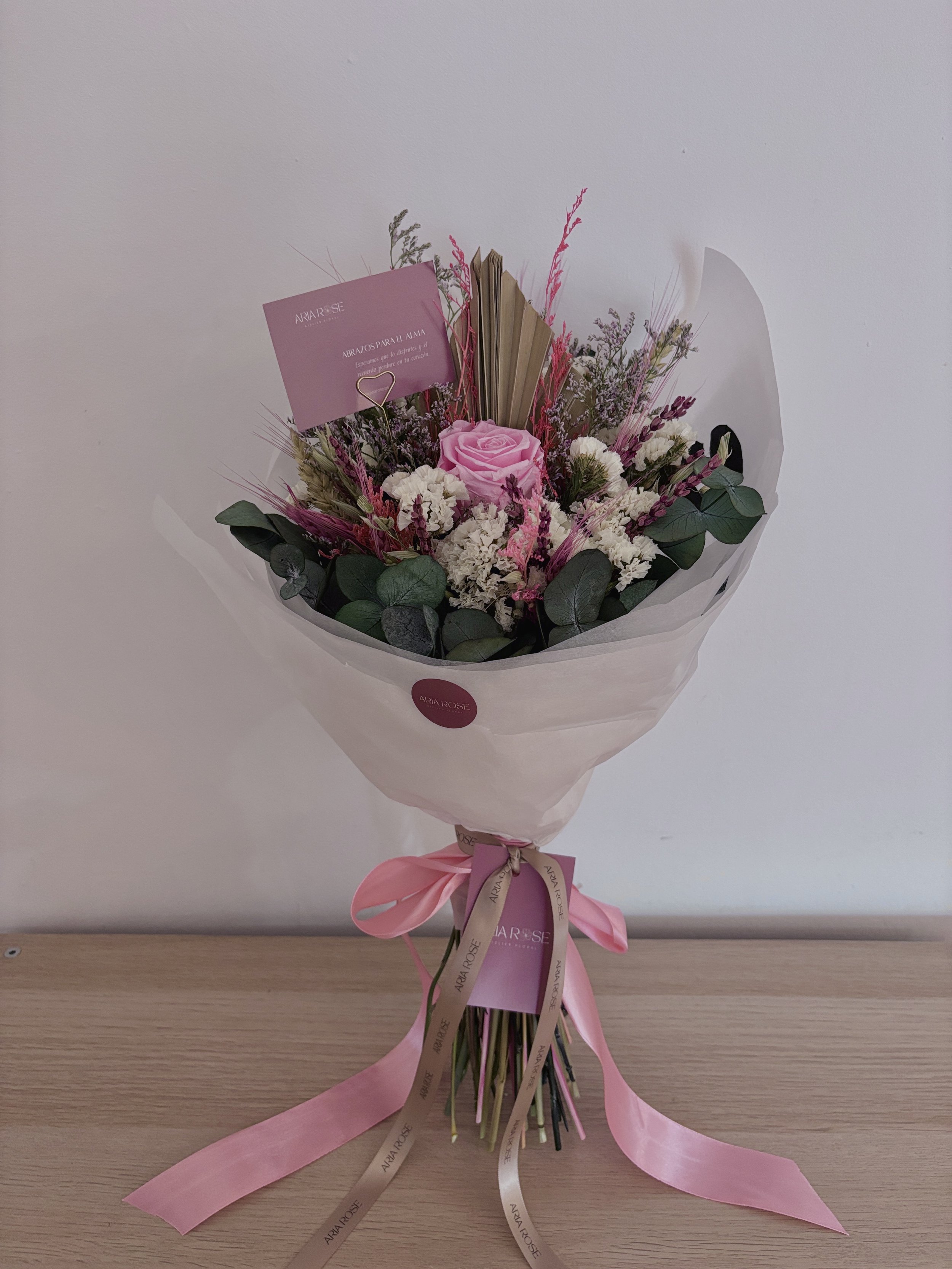 Ramo de flores con rosas rosas, flores blancas, hojas verdes, y decoraciones con cintas en color rosa y beige, envuelto en papel transparente y una etiqueta que dice 'AIRA ROSE'.