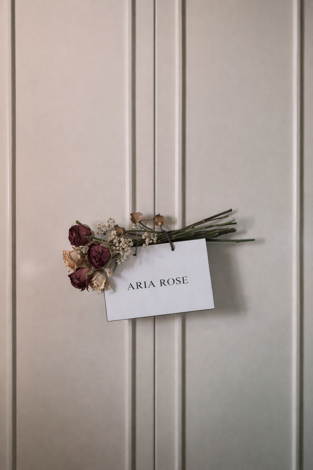 Ramo de rosas secas con una etiqueta que dice 'ARIA ROSE', colgado en una puerta de color gris claro.