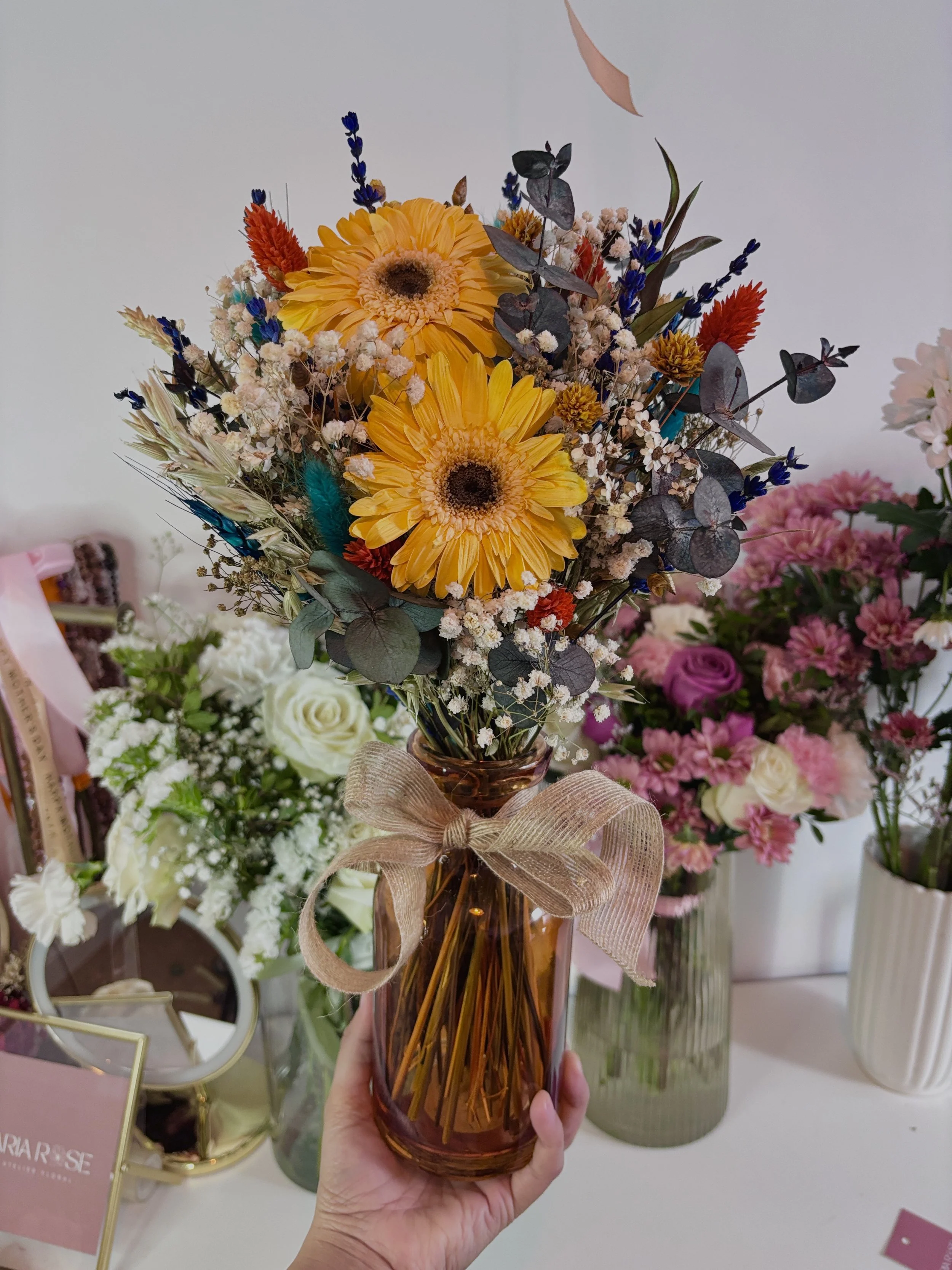 Ramo de flores en jarrón ámbar, flores amarillas, blancas, rosas, lilas y verdes con lazos de tela, en un ambiente floral decorativo.