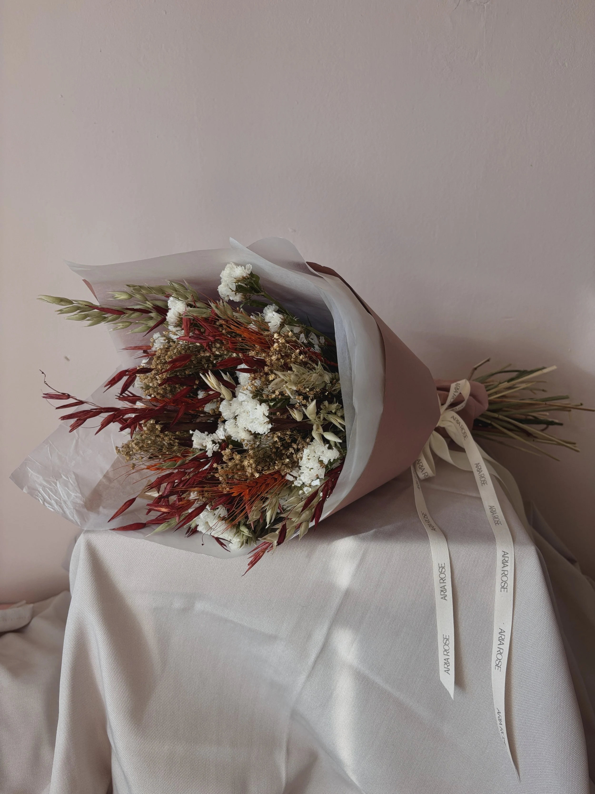 Ramo de flores secas y blancas envuelto en papel y cinta con la inscripción 'ARÍA ROSE', colocado sobre una mesa cubierta con tela blanca.