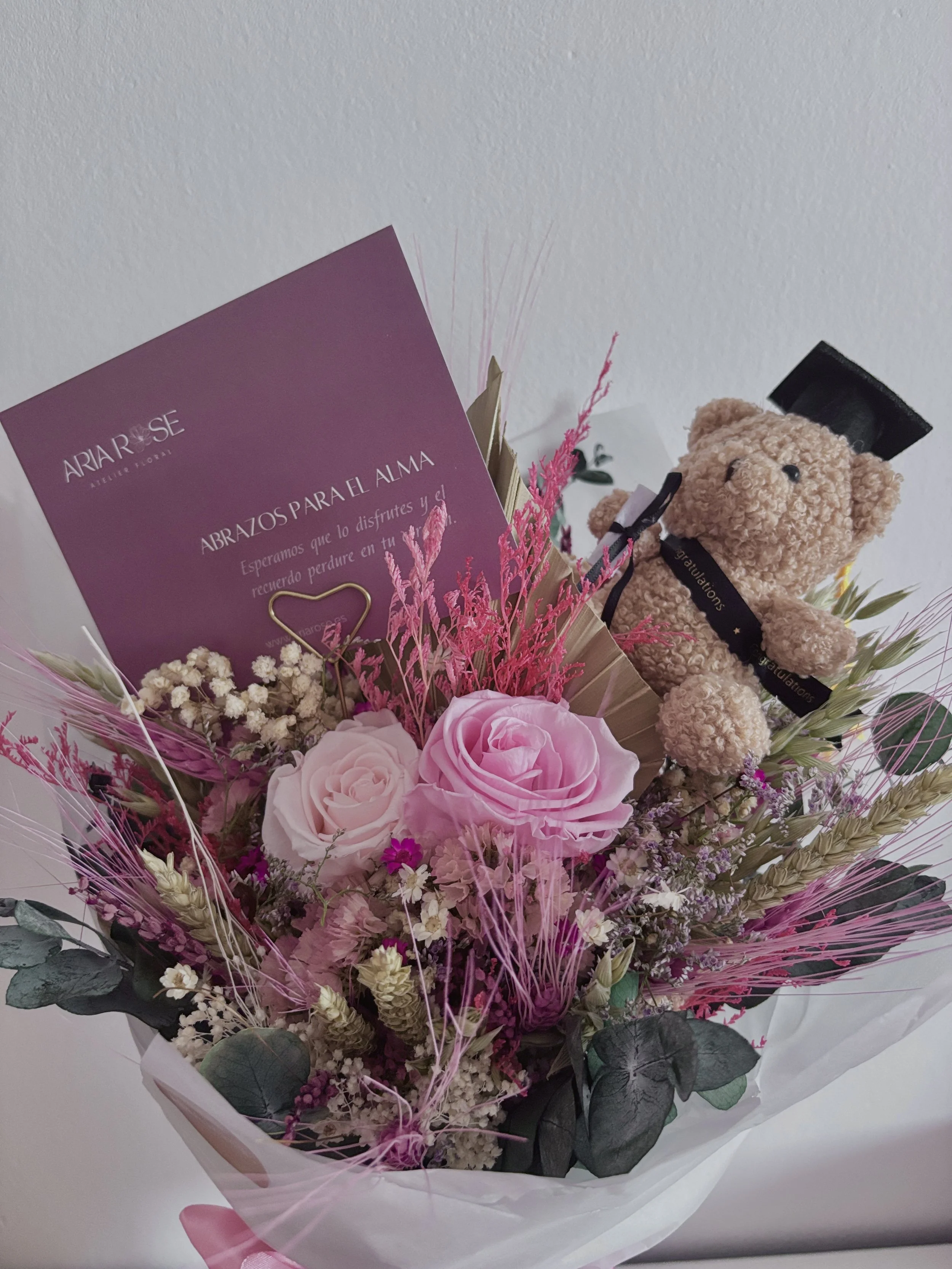 Arreglo de flores con rosas rosadas, flores blancas y secas en tonos rosados, acompañado de un osito de peluche con birrete de graduación y tarjeta con mensaje en español.