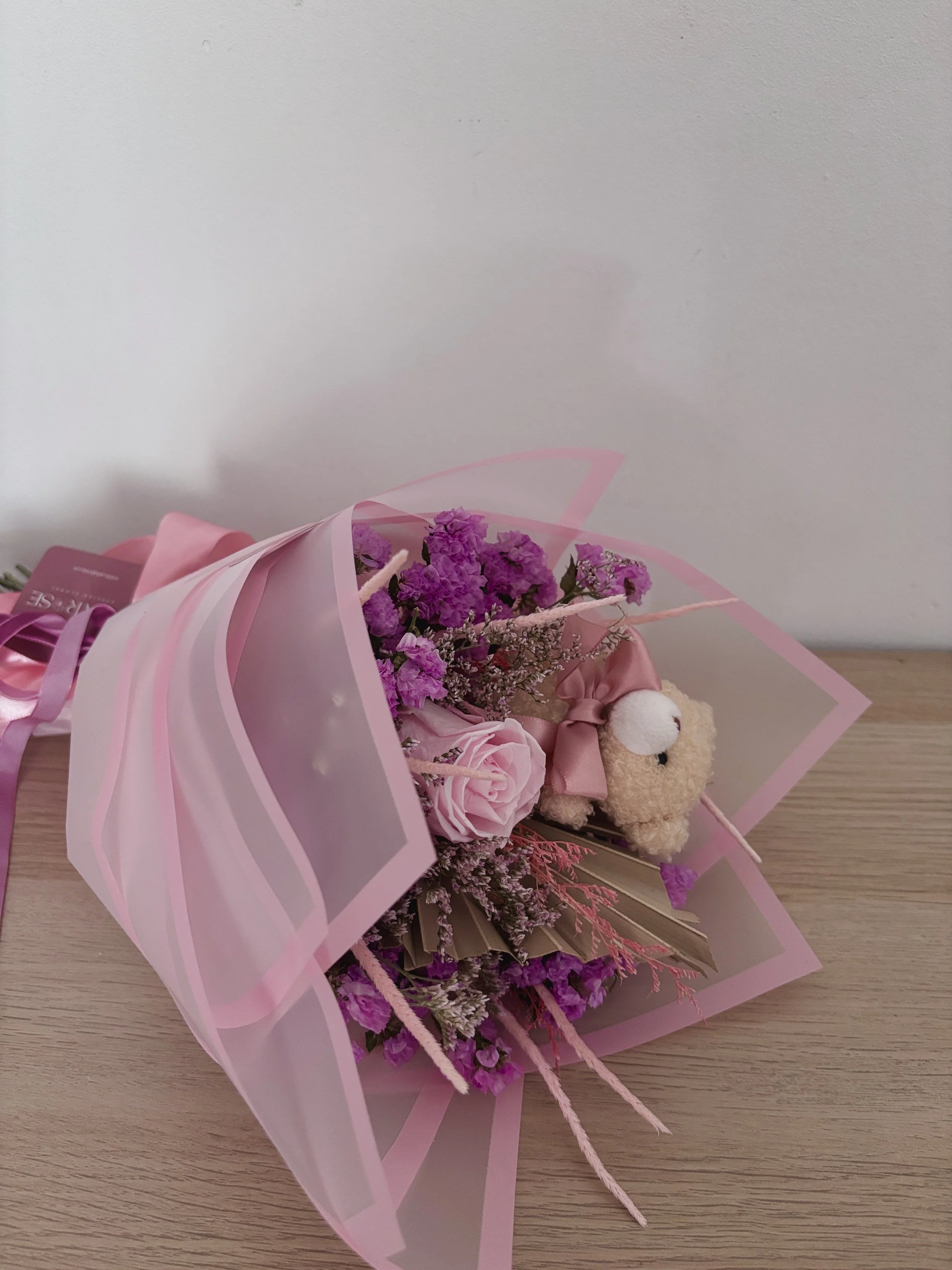 Ramo de flores con rosas y flores moradas, envuelto en papel rosado y decorado con una pequeña oveja de peluche
