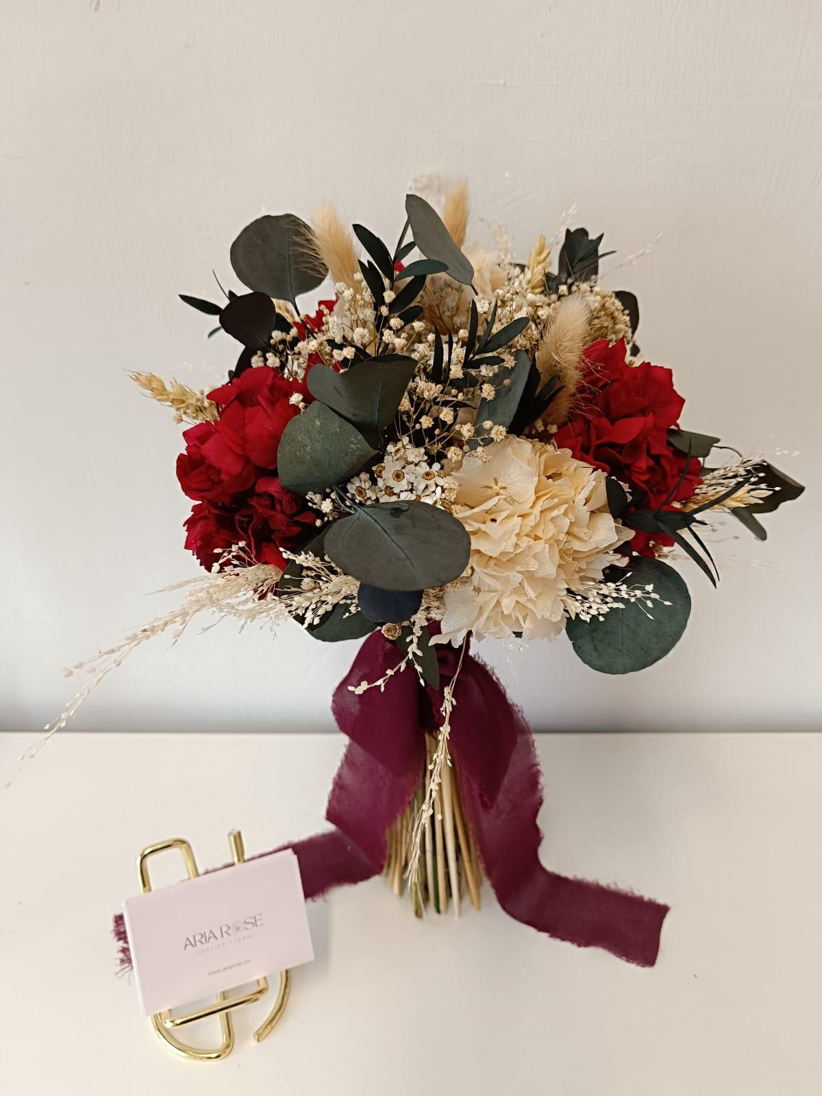 Ramo de flores con rosas rojas, flores blancas, follaje verde y decoraciones dried floral, sujeto con cinta de terciopelo morada y acompañado de una tarjeta y ganchos dorados.
