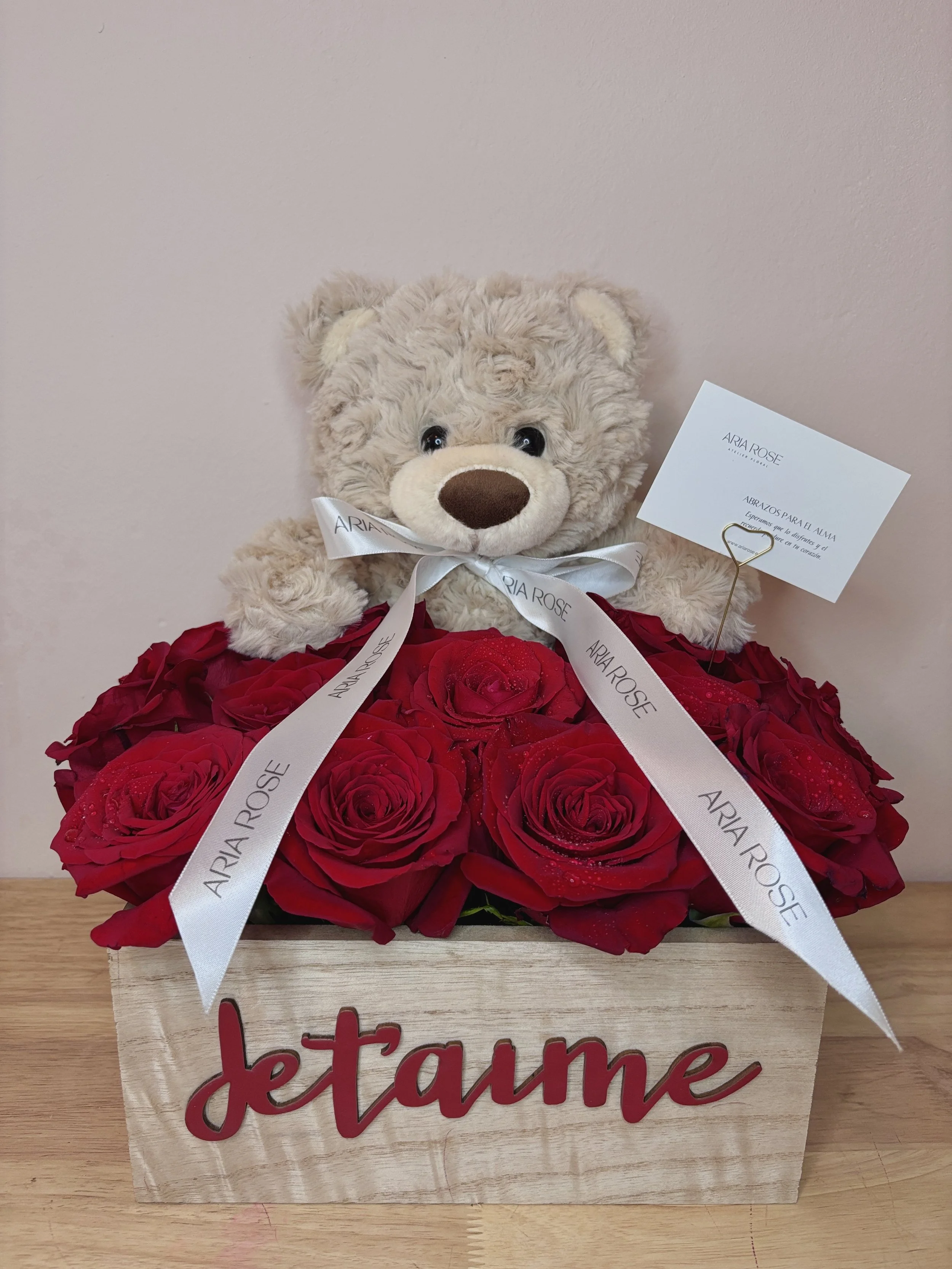 Un arreglo de flores rojas en una caja de madera con la palabra 'te amo' en letras rojas, acompañado de un oso de peluche y una tarjeta con mensaje, decorado con una cinta que dice 'ARIA ROSE'.