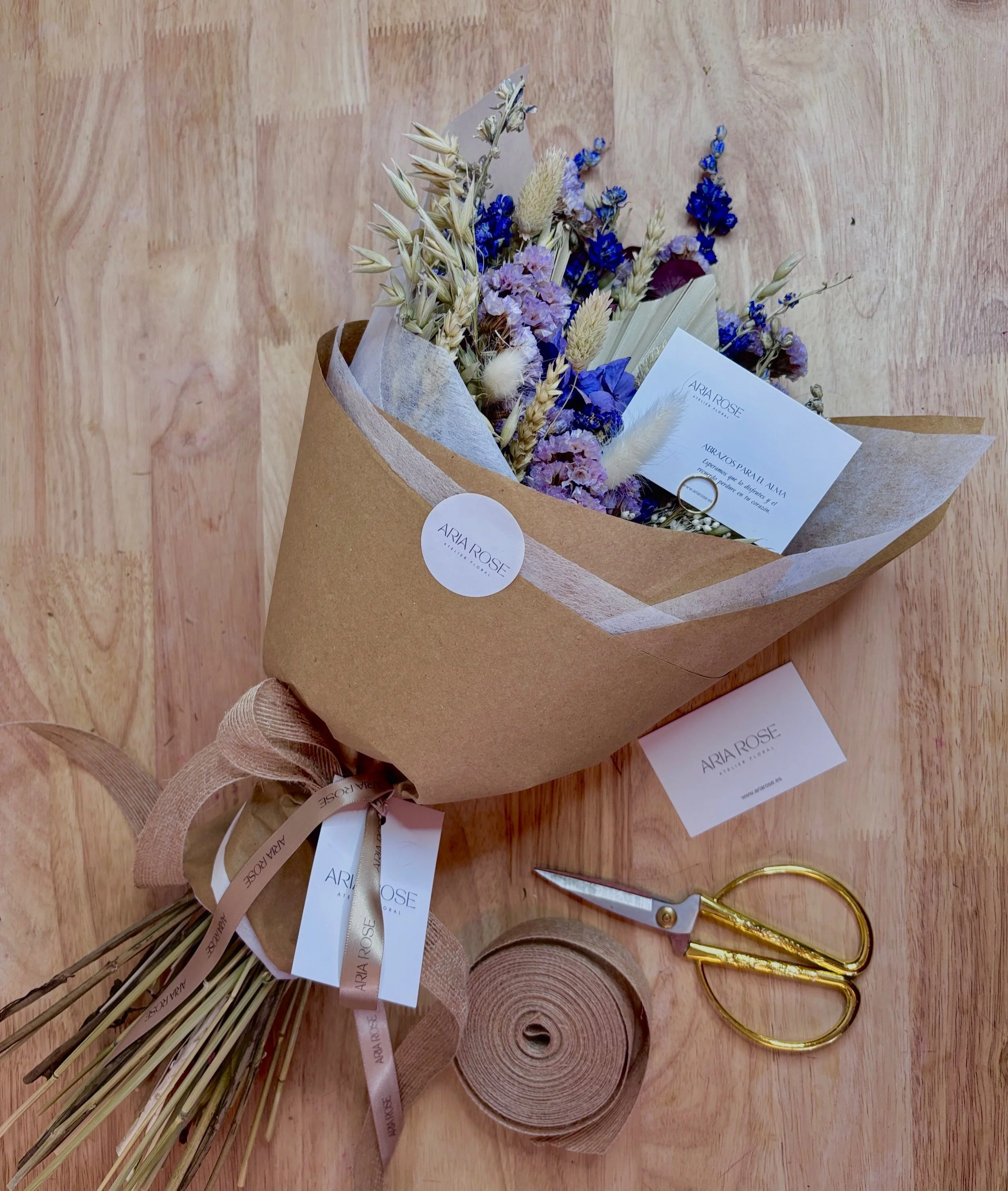 Ramo de flores secas en papel marrón, acompañado de tijeras doradas y cinta decorativa en un suelo de madera.