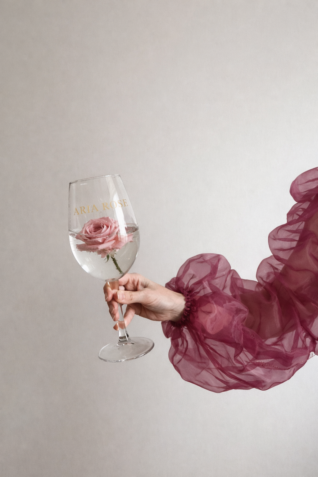 Vaso de vidrio con rosa en agua, con la inscripción 'ARIA ROSE', sostenido por una mano con manga de tul rosa rizado y transparente.