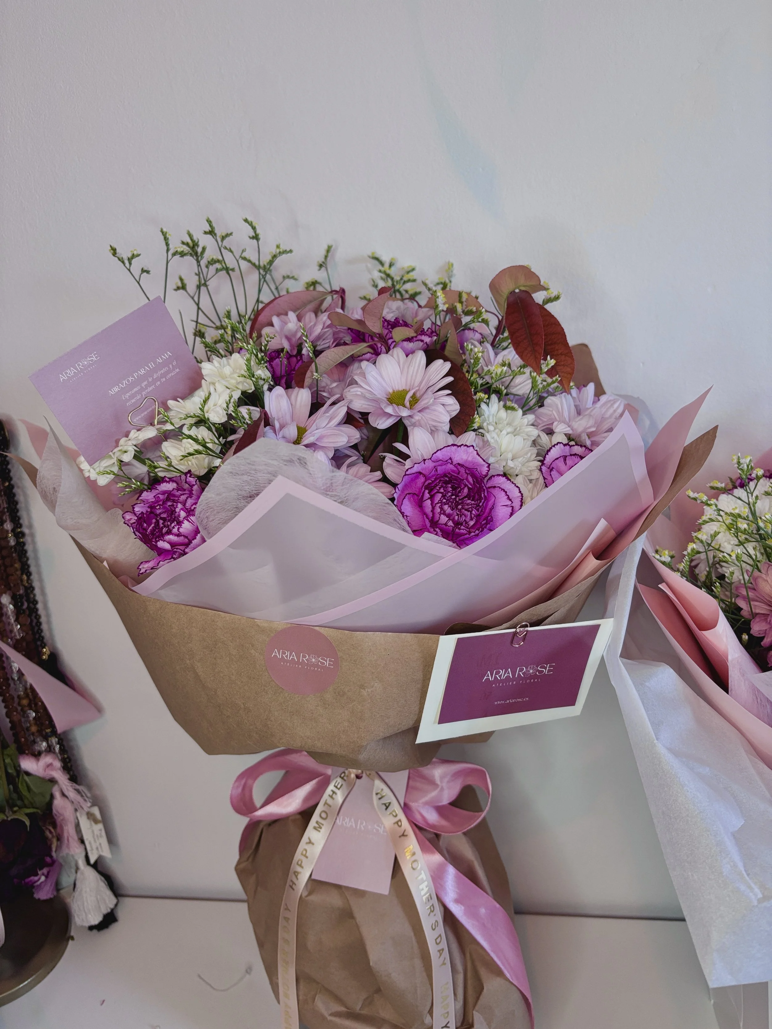 Ramo de flores con rosas, margaritas y otras flores en tonos rosa y morado, envuelto en papel decorativo y lazo rosa, con una tarjeta que dice 'ARIA ROSE'.
