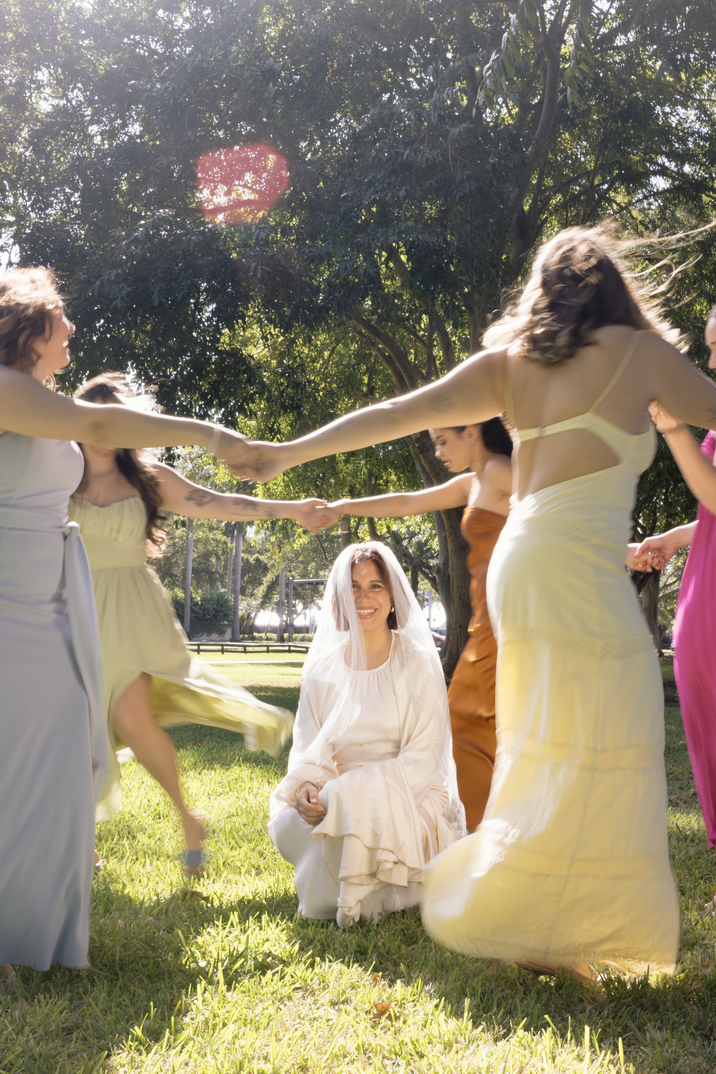 Bridesmaids circle bride under trees, playful moment in documentary style wedding photography