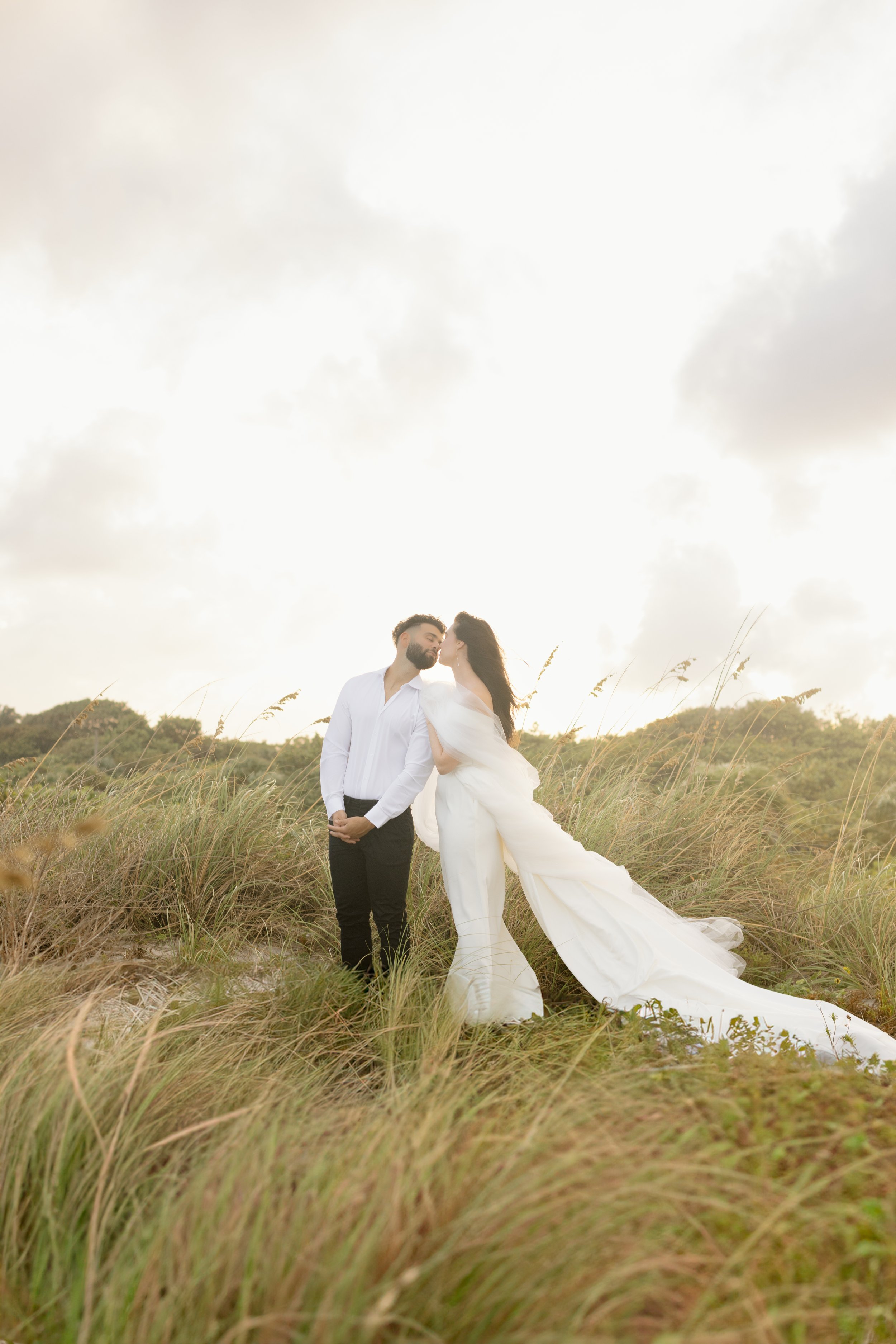 Groom kisses bride in grassy field, golden hour in documentary style wedding photography