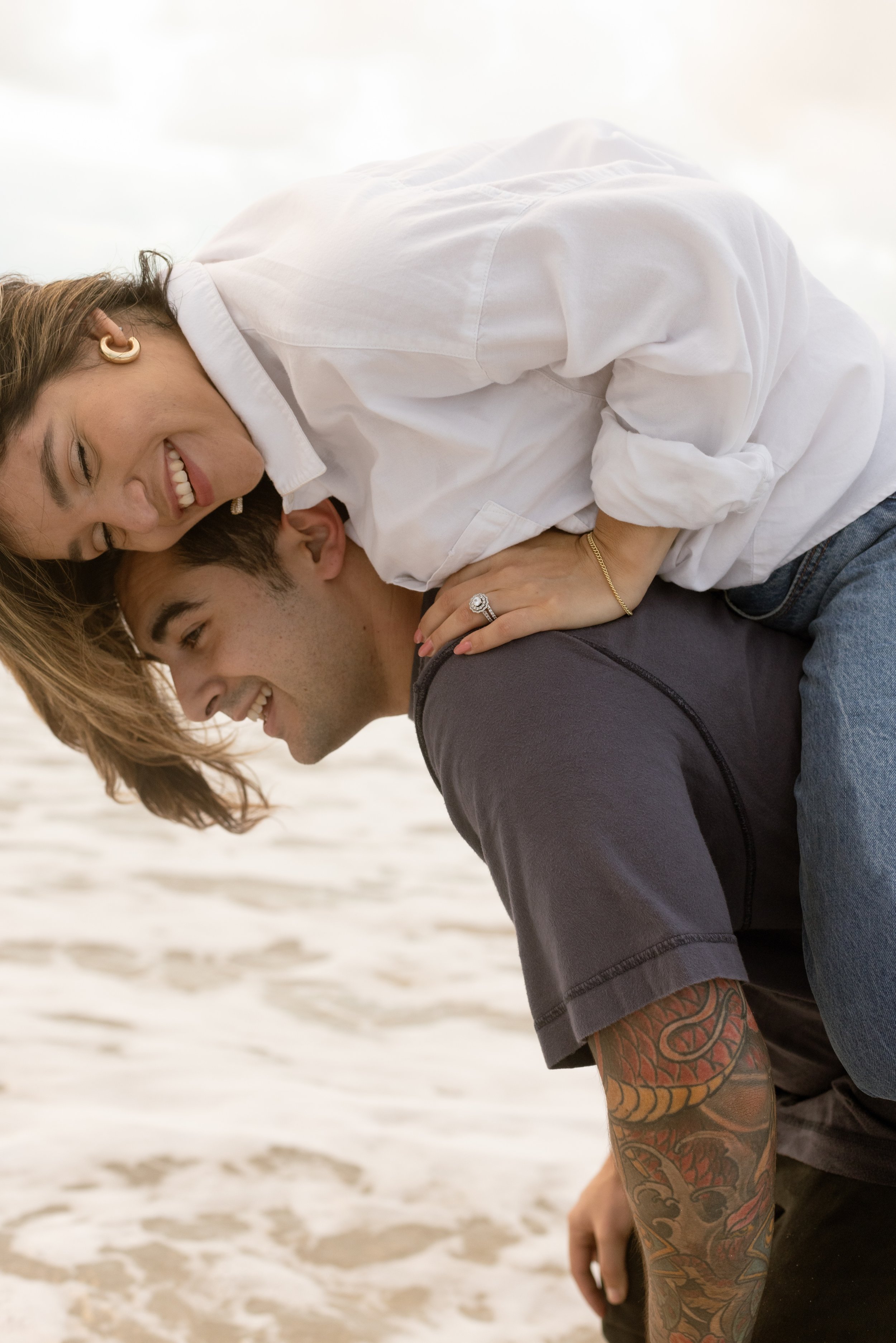Bride laughs riding piggyback on groom, beach fun with documentary wedding photographer