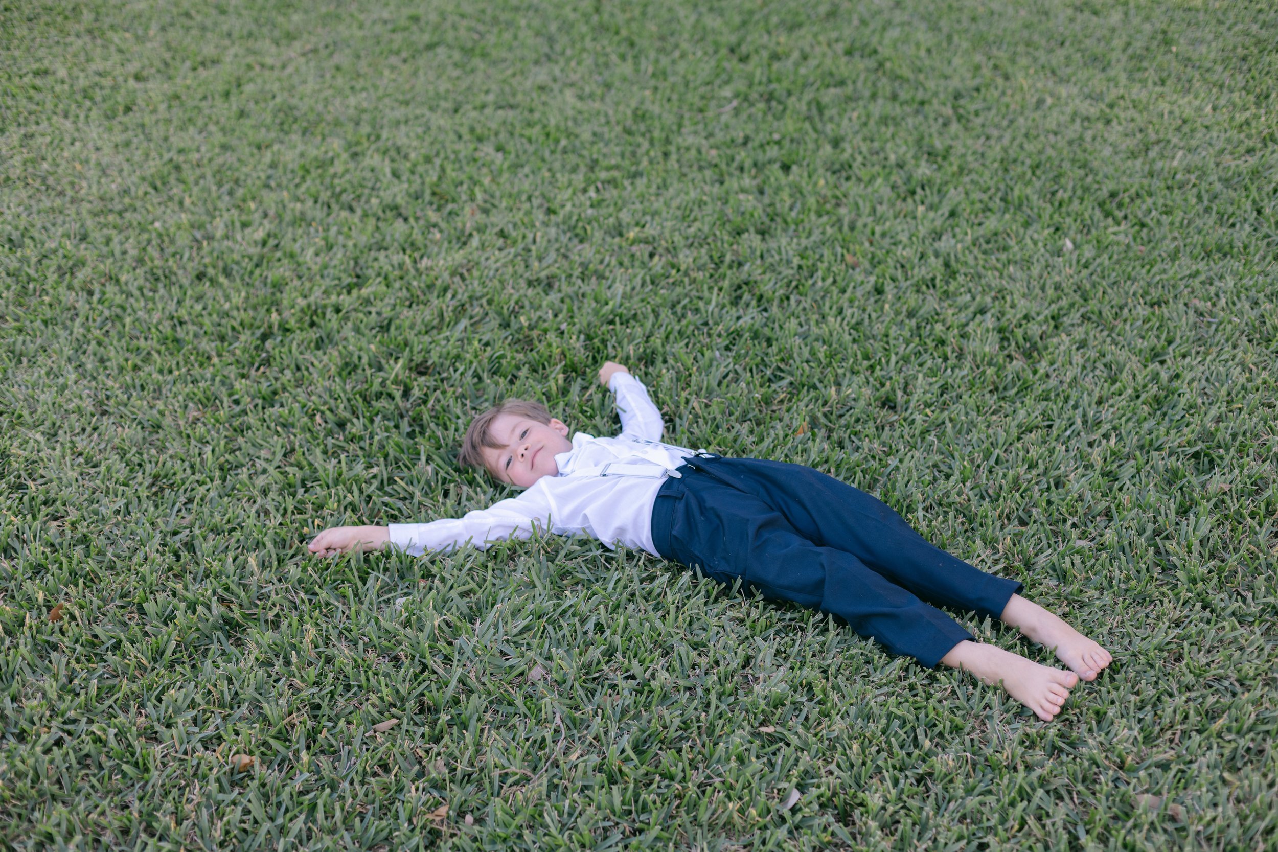 Young boy laying on green grass, playful wedding moment in documentary style