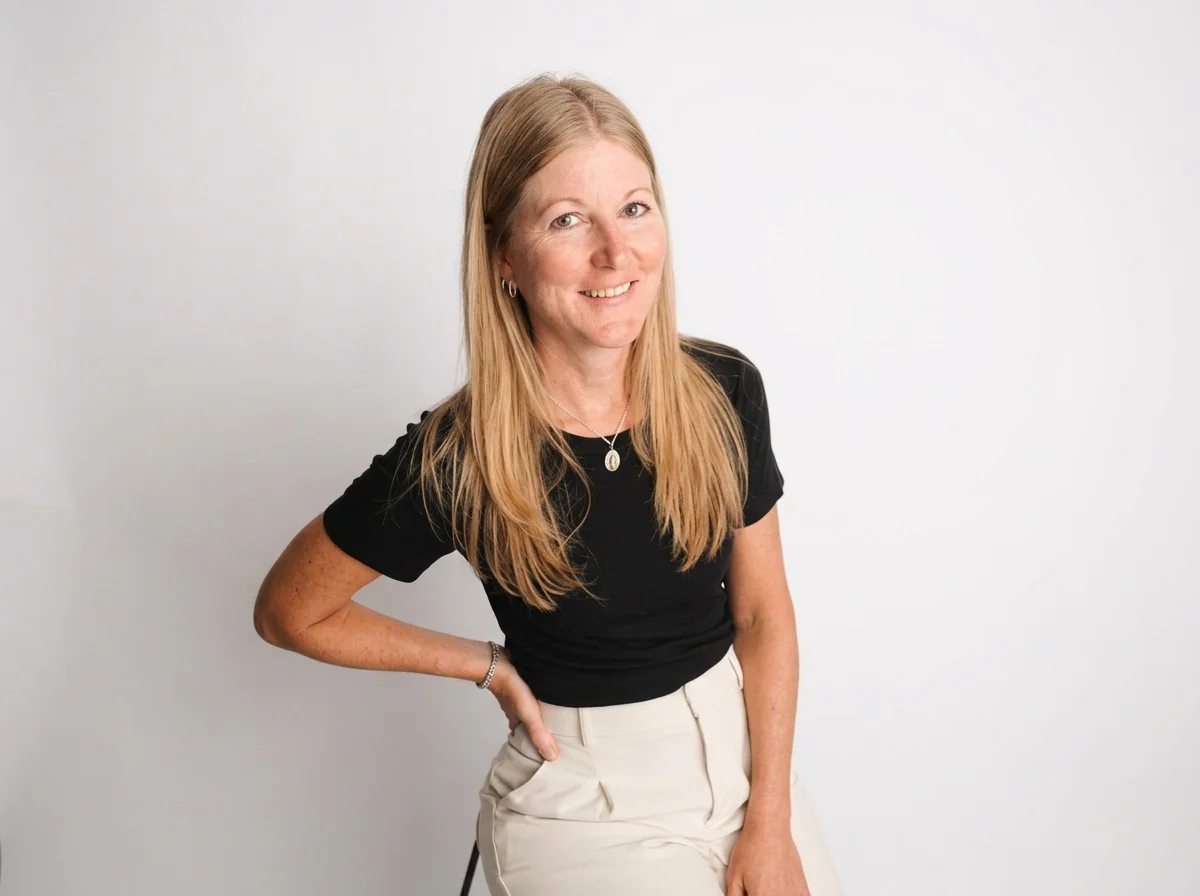 Smiling blonde woman in a black shirt and cream pants posing against a white background