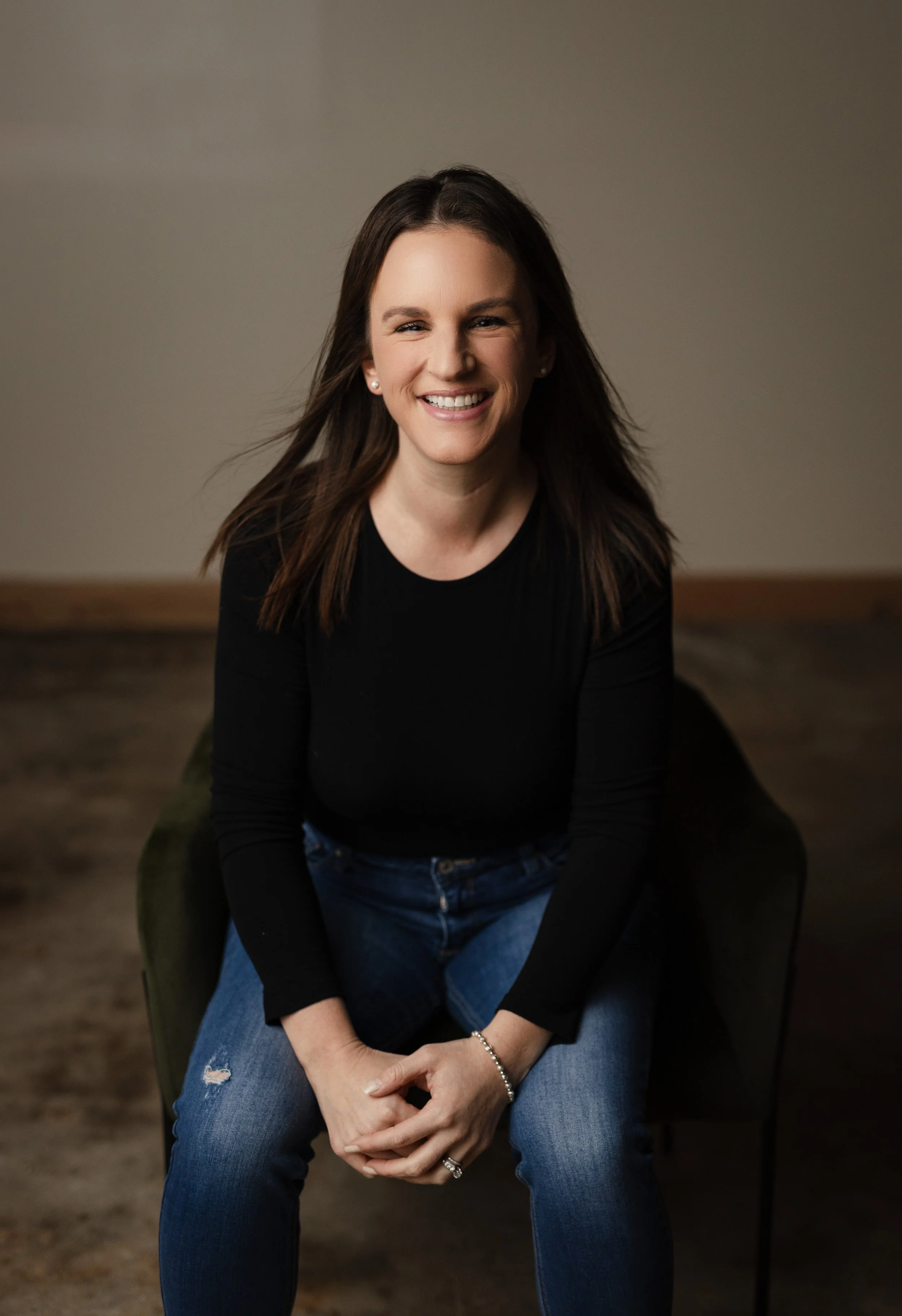 Smiling woman with long brown hair seated indoors, wearing a black top and blue jeans.
