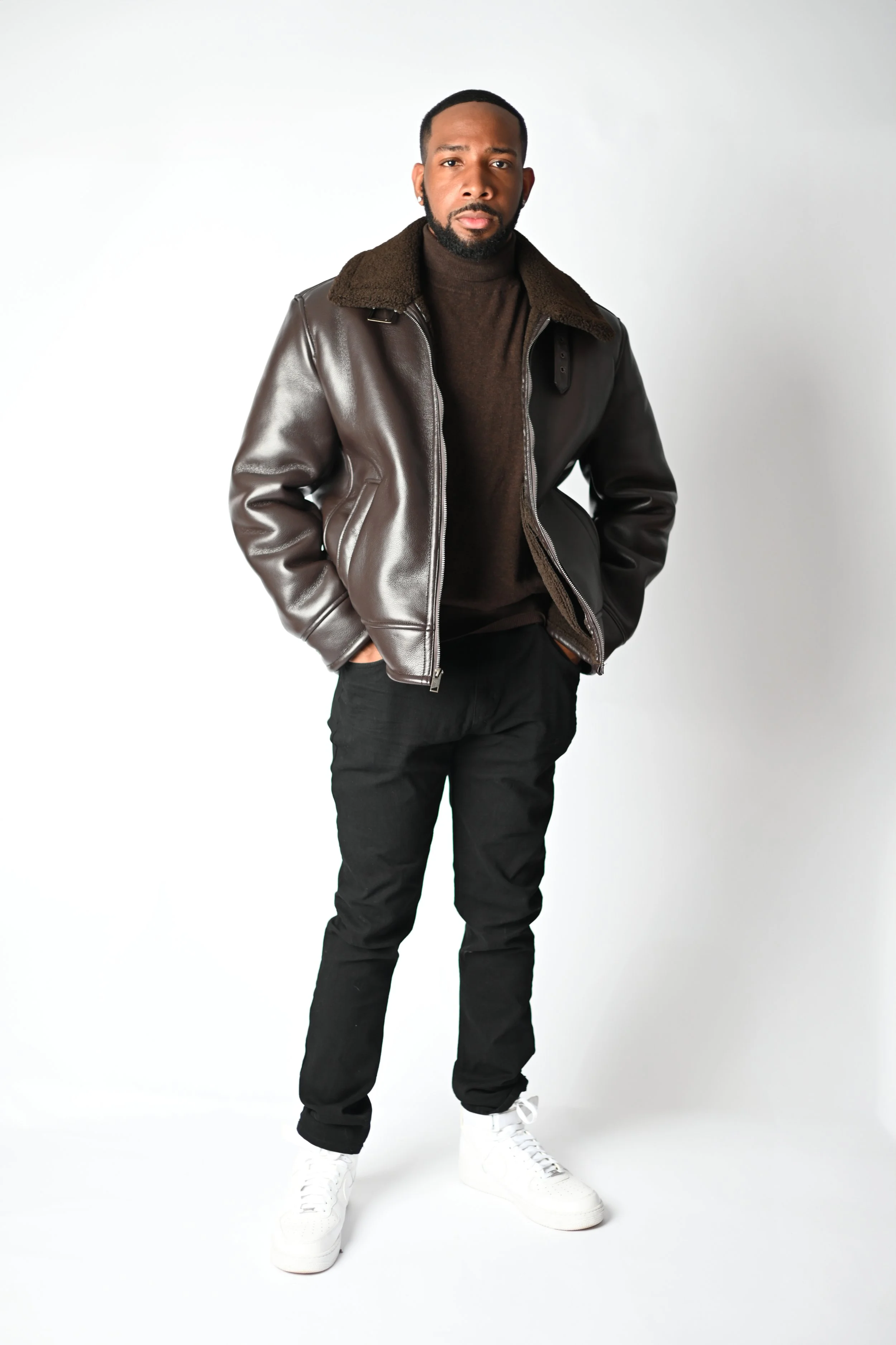 A young man with a beard and short hair standing against a plain white background. He is wearing a dark brown leather jacket with a shearling collar, a brown turtleneck. Pittsburgh actor headshots photoshoot Wolfsbora Media