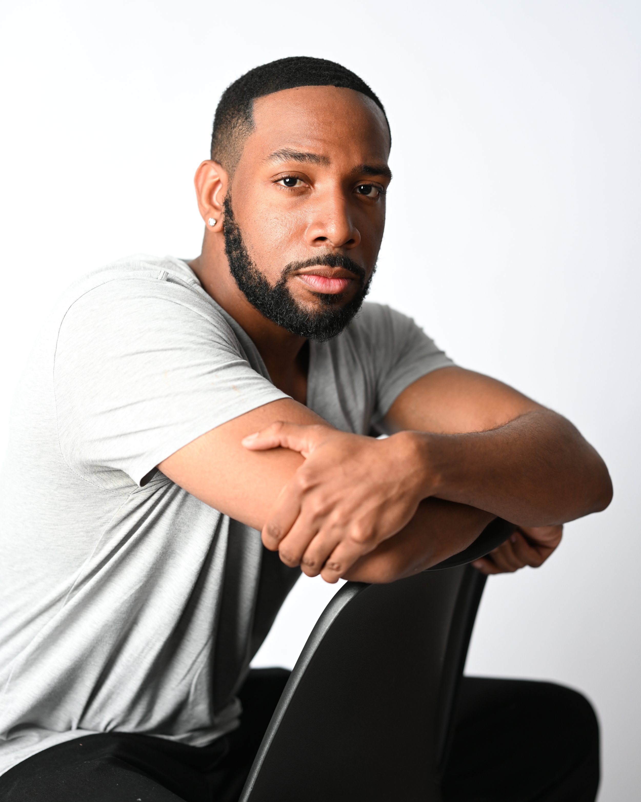 A man with a beard and earrings, wearing a light gray t-shirt, sits with his arms crossed over black chairs against a white background. Pittsburgh photoshoot portrait headshots actor photoshoot photography Wolfsbora Media