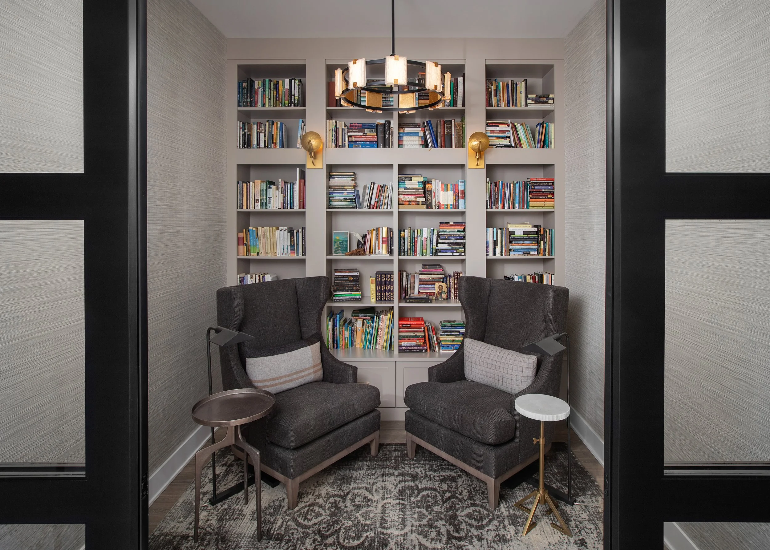 Cozy reading nook in a DEV Partners development in Pella, Iowa, featuring elegant built-in bookshelves filled with books, two stylish armchairs, and modern wall sconces, highlighting sophisticated living spaces ideal for real estate investment.