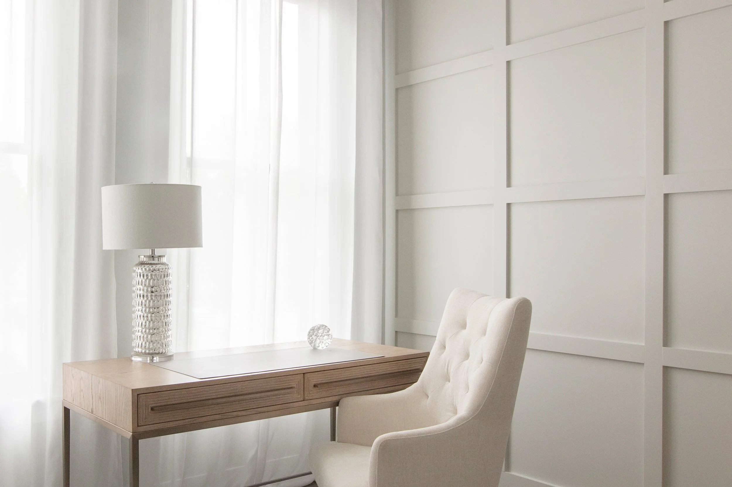 Serene study corner in a Pella, Iowa property by DEV Partners, featuring a stylish desk, a comfortable tufted chair, and a modern lamp, set against elegant white panel walls, exemplifying sophisticated living spaces for those investing in real estate