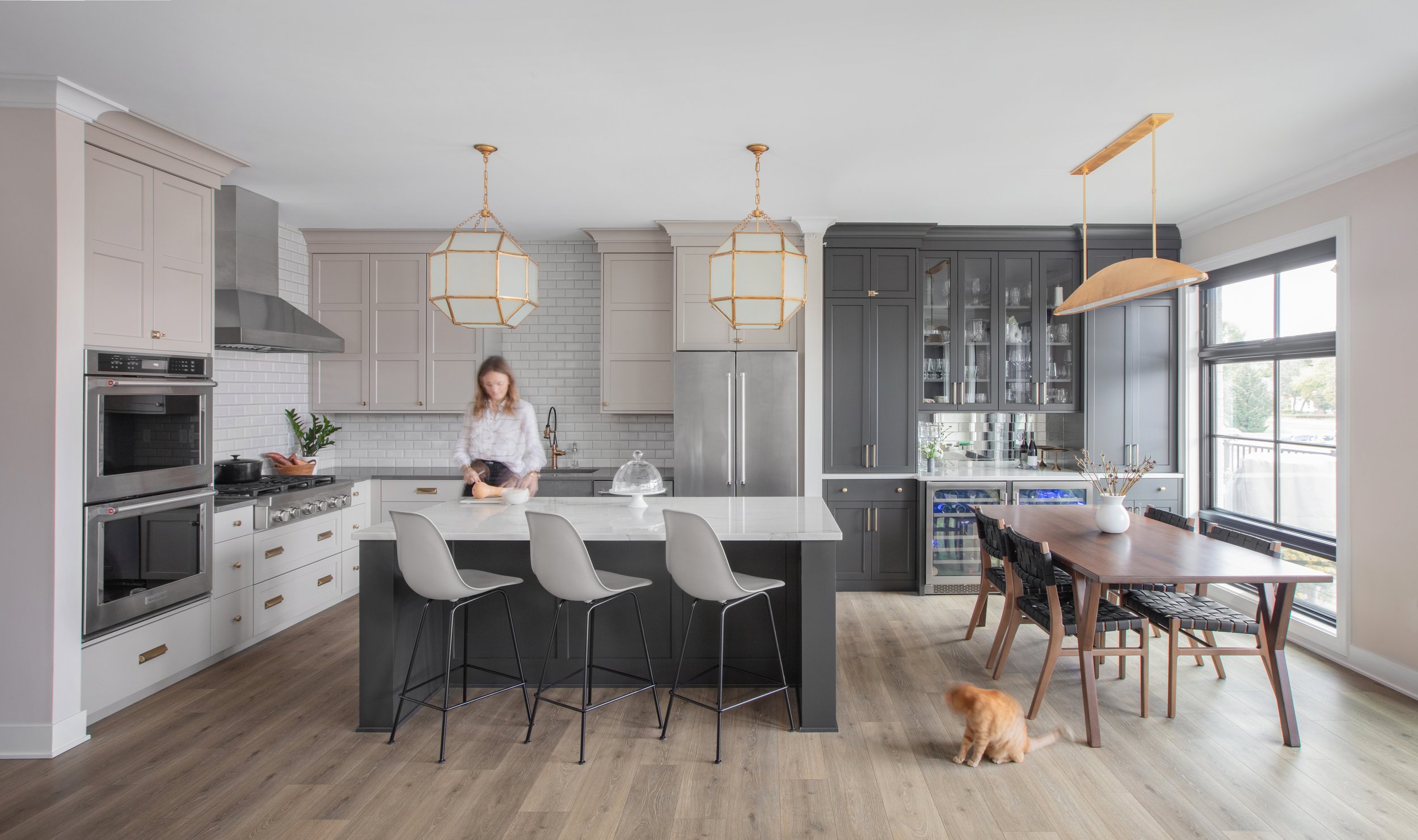 Elegant interior of a DEV Partners investment property showcasing a spacious modern kitchen with gray cabinetry and white tile backsplash, a large dining area a wooden table, and gold lighting fixtures. A woman prepares food at the kitchen island
